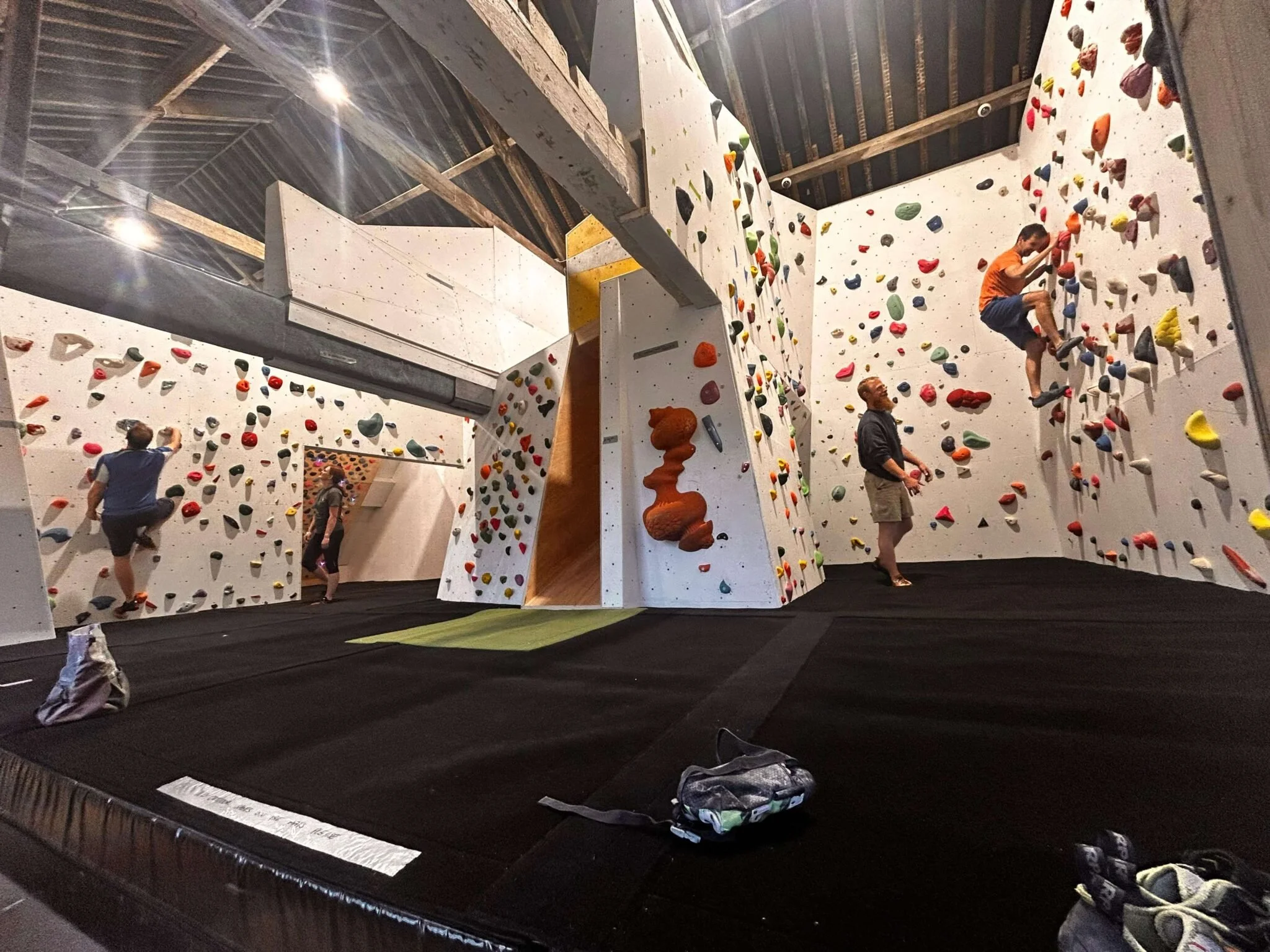 Indoor rock climbing gym with colorful climbing holds, climbers, and a trainer, featuring a high ceiling with exposed beams.