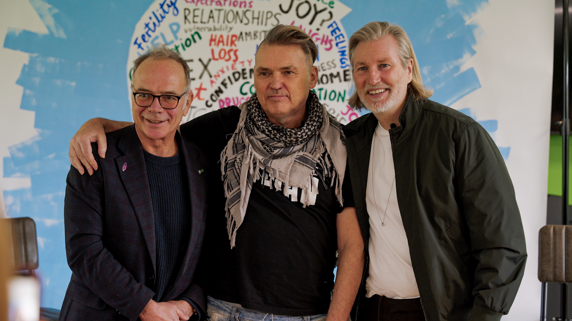 Three men standing close together with their arms around each other, smiling for the camera, in front of a colorful wall with words related to relationships and feelings.