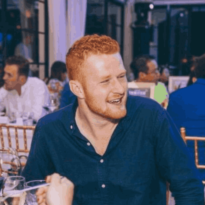 A young man with red hair and a beard wearing a navy blue shirt, smiling and laughing at a social gathering indoors.