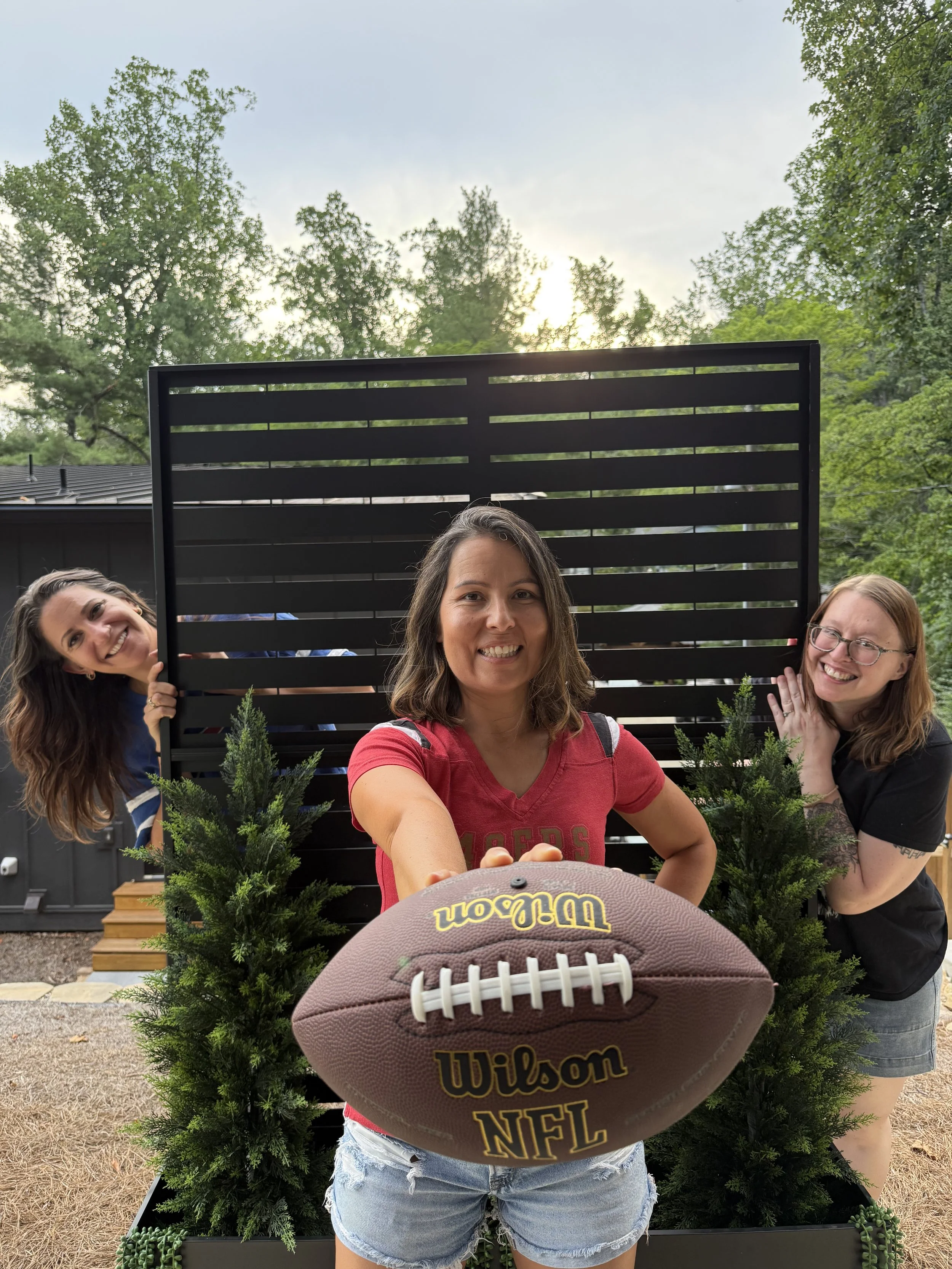 Woman standing front of a fence holding an NFL football. Two women peaking out from behind the fence.