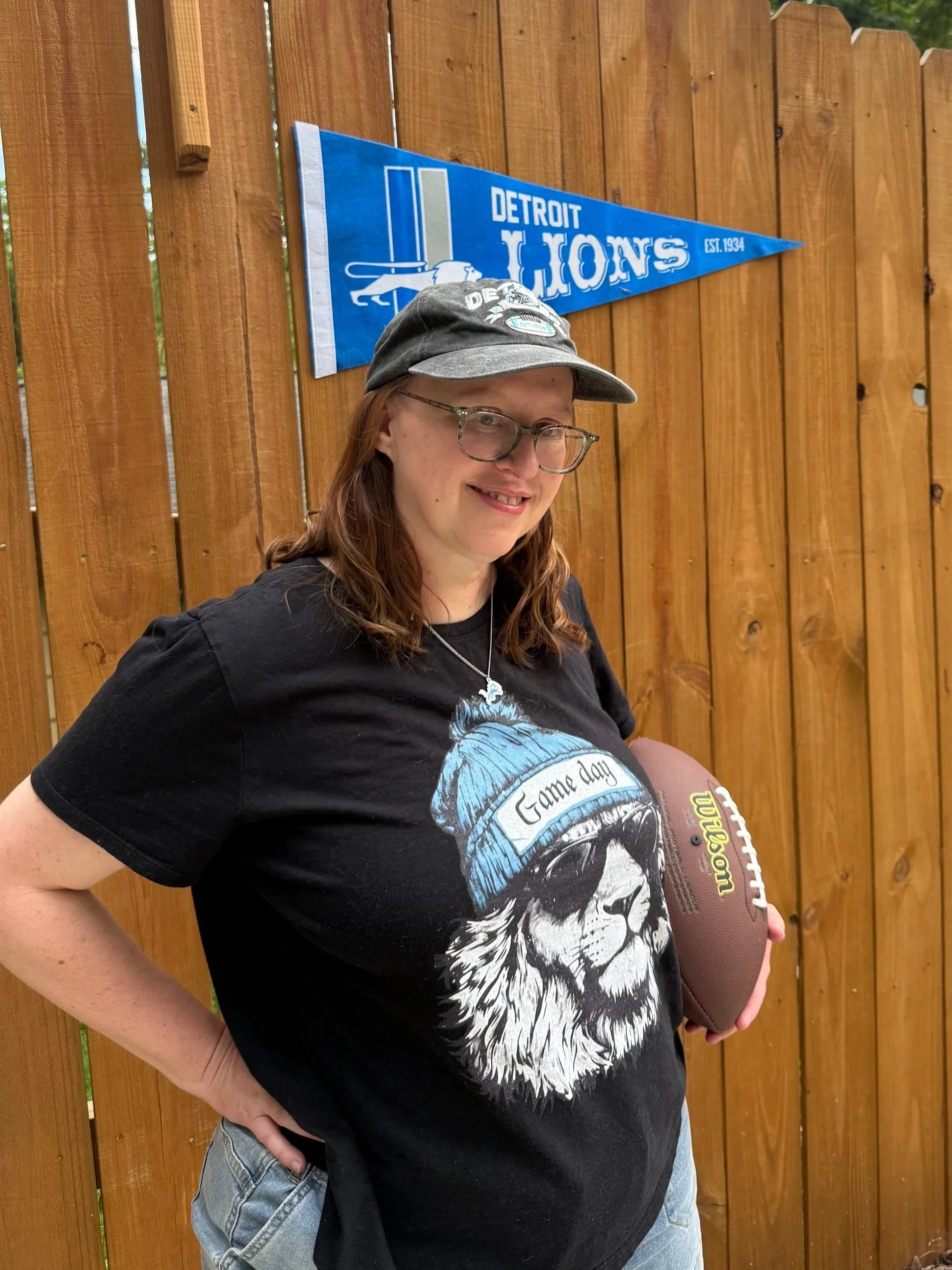 Red haired woman with glasses wearing a Detroit lions tshirt and hat holding a football standing in front of a Detroit Lions pennant.