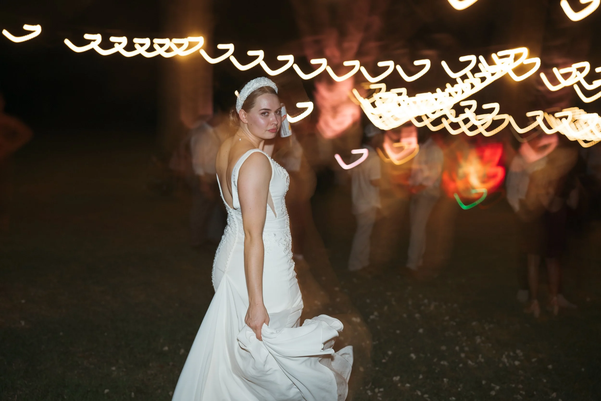 Bride walking back to the dance floor with a beer in hand, playfully swinging the tail of her gown as she dances with colorful lights trail behind her using flash drag at Amelia grove in New Bern NC| fun and artistic wedding reception photography
