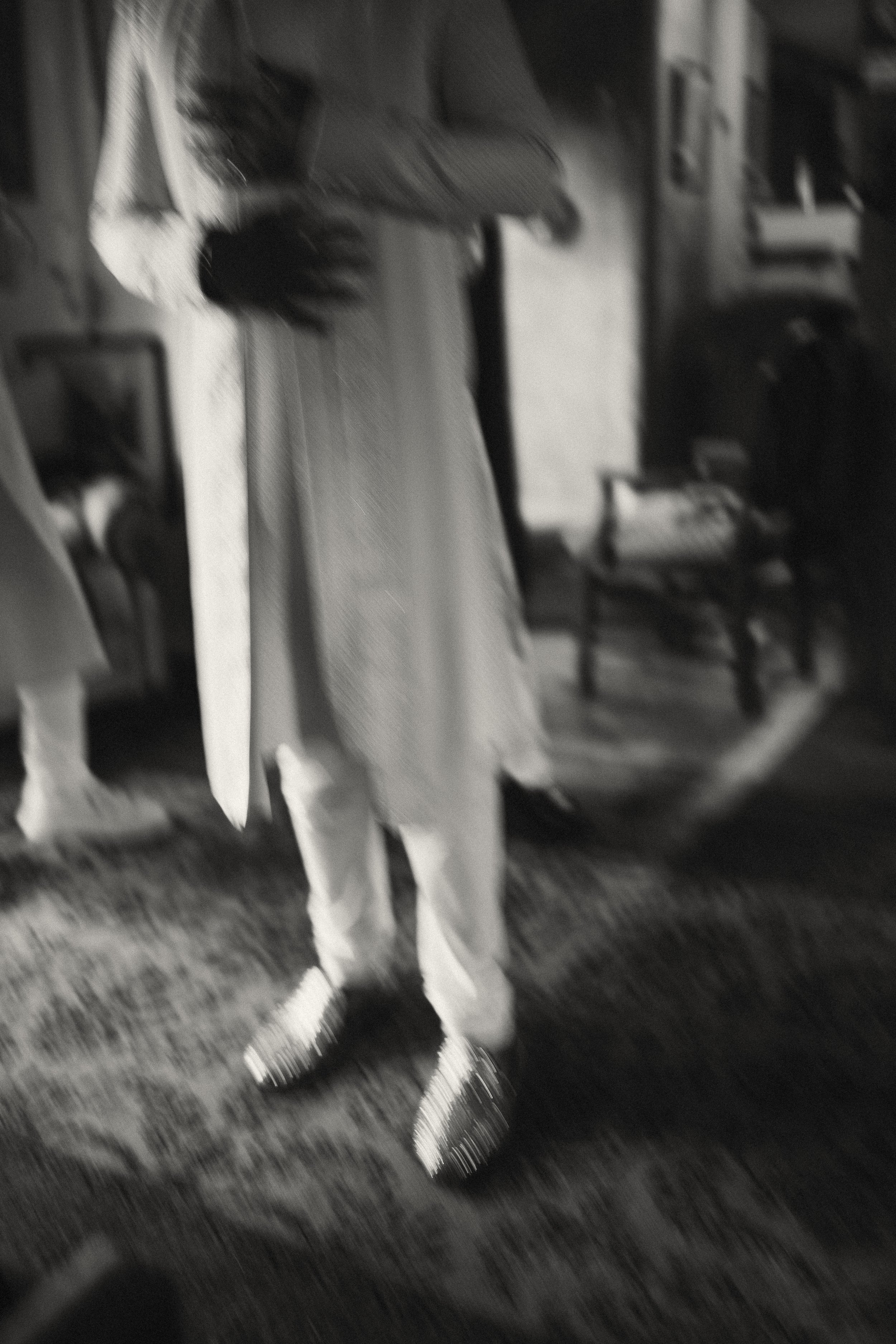 this is a blurry black and white photo of the groom's hands while he straightens out his beautiful traditional Indian garments. the sparkle on his shoes make the photo more visually interesting.