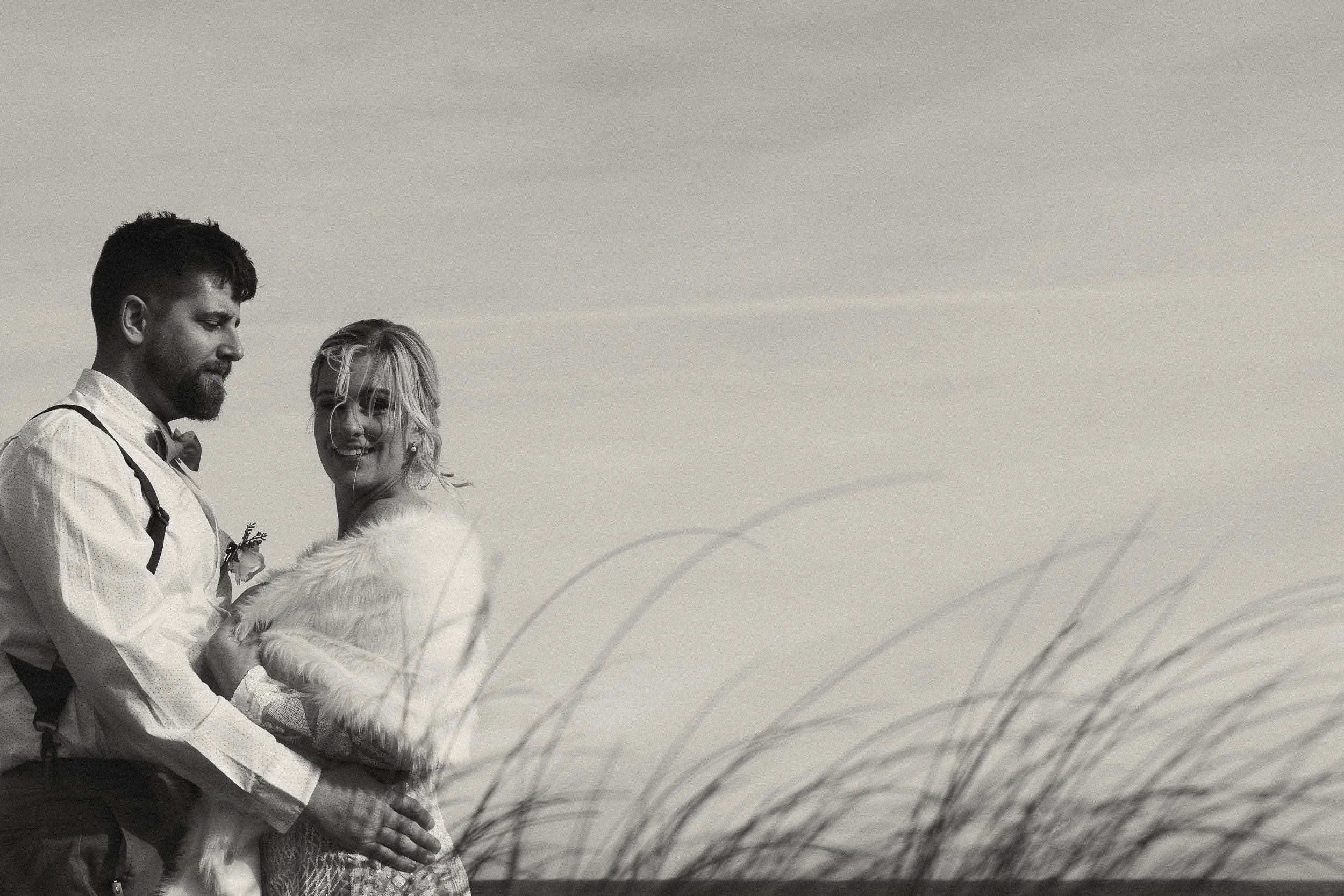 Black and white photo of a winter elopement at Kure Beach, NC, with the bride in a sparkly gown and fur wrap standing by the ocean rocks | emotional and timeless oceanside wedding photography