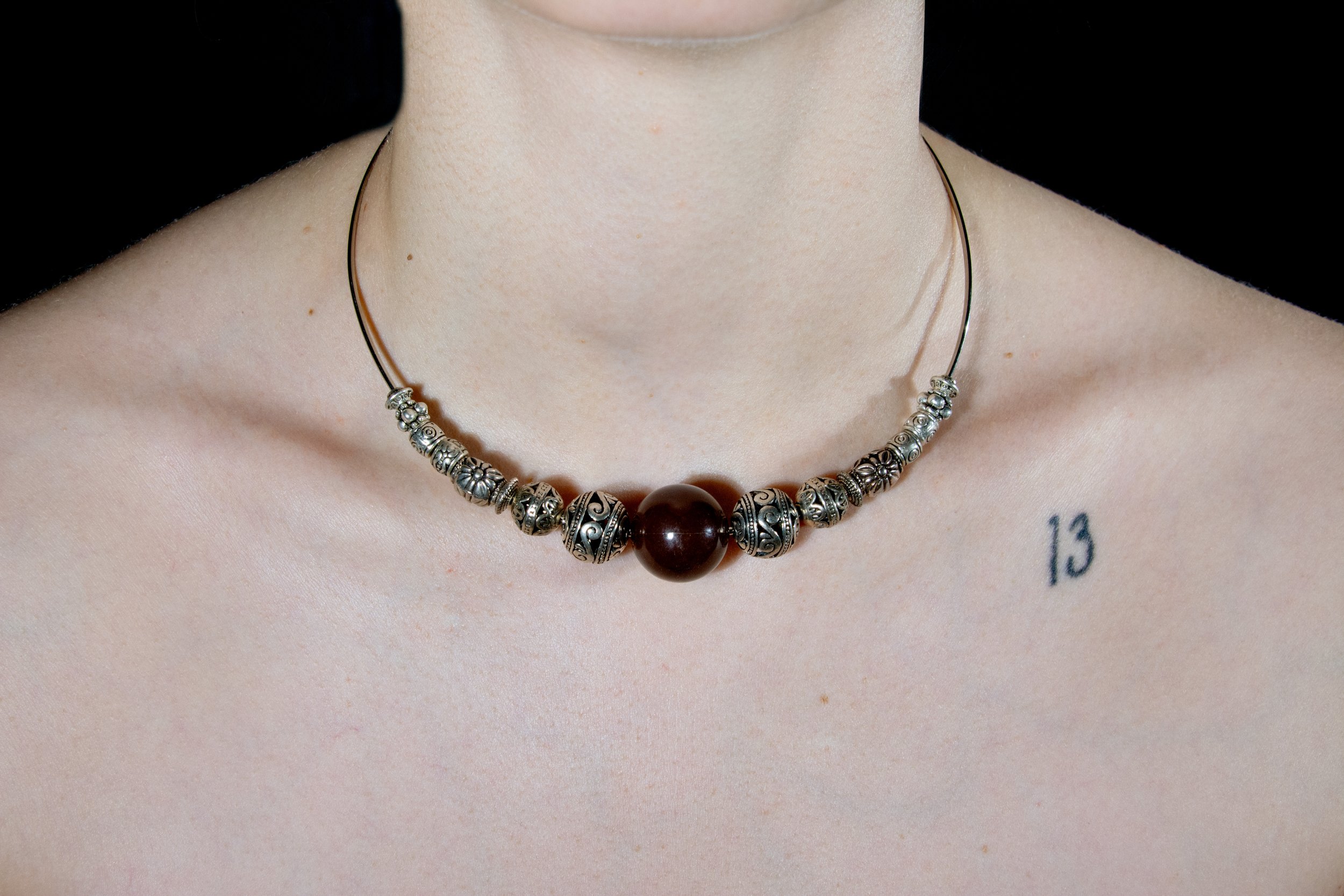 Model's neck and chest area pictured wearing steel wire choker necklace with varied silver beads and large brown statement bead on black background