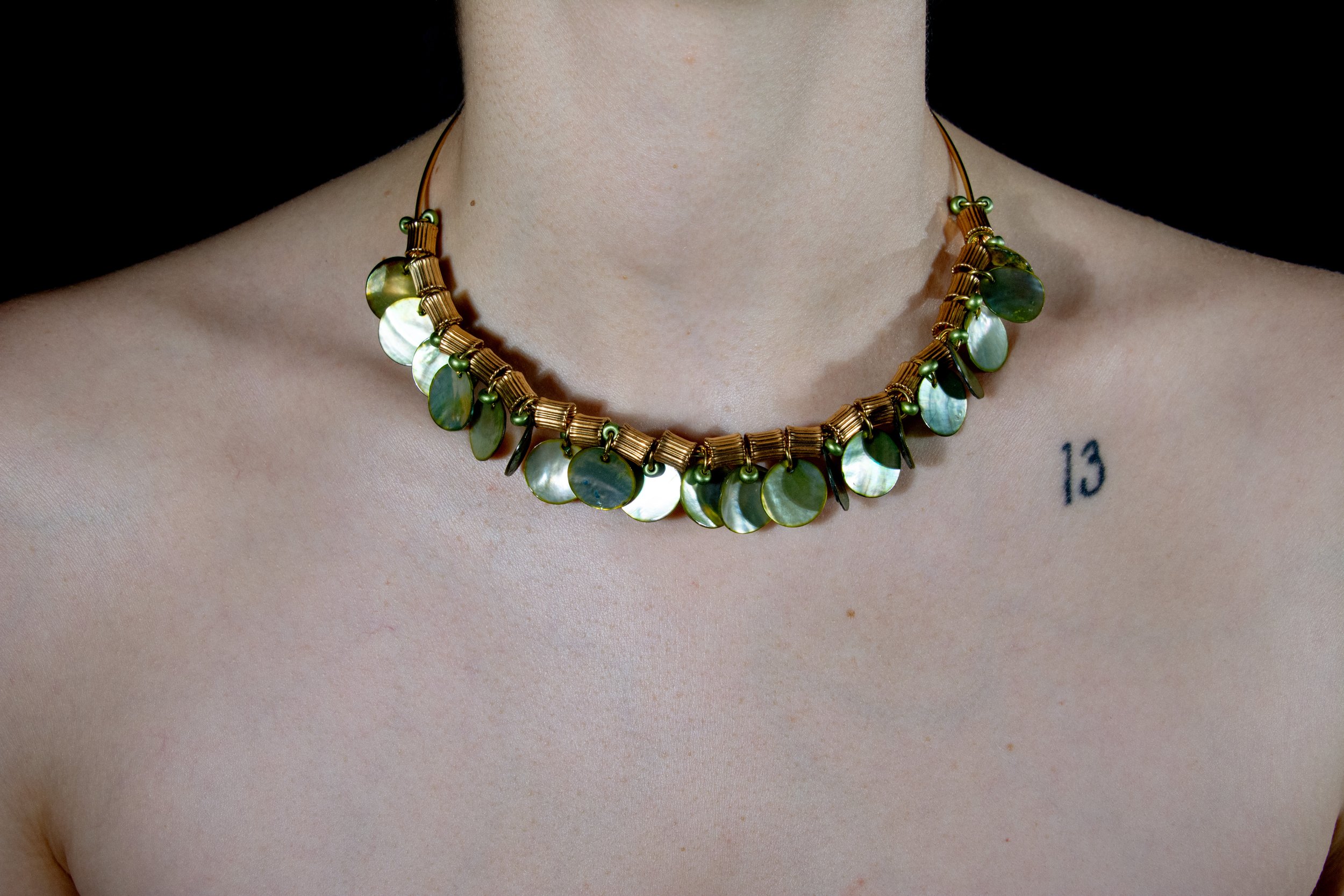 Model's neck and chest area pictured wearing steel wire choker necklace with varied ornate gold beads and thin iridescent green shell charms on black background