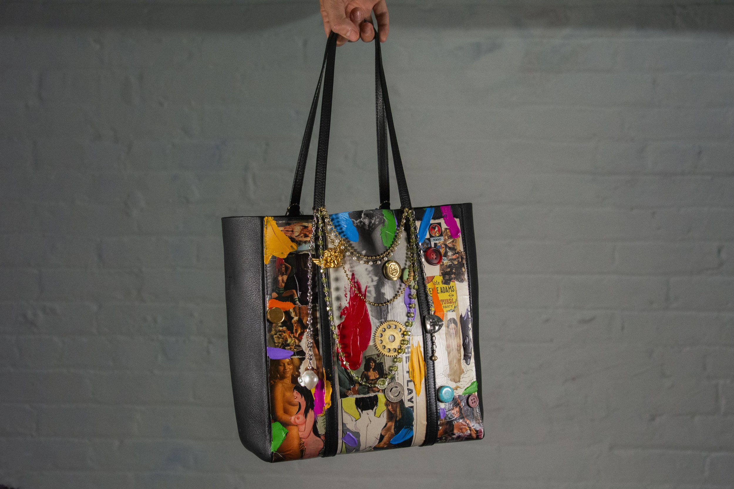 Backside of painted, collaged, and sealed Balenciaga black purse with colorful button additions, various charms, beads & hardware being held by it's straps against a white brick wall