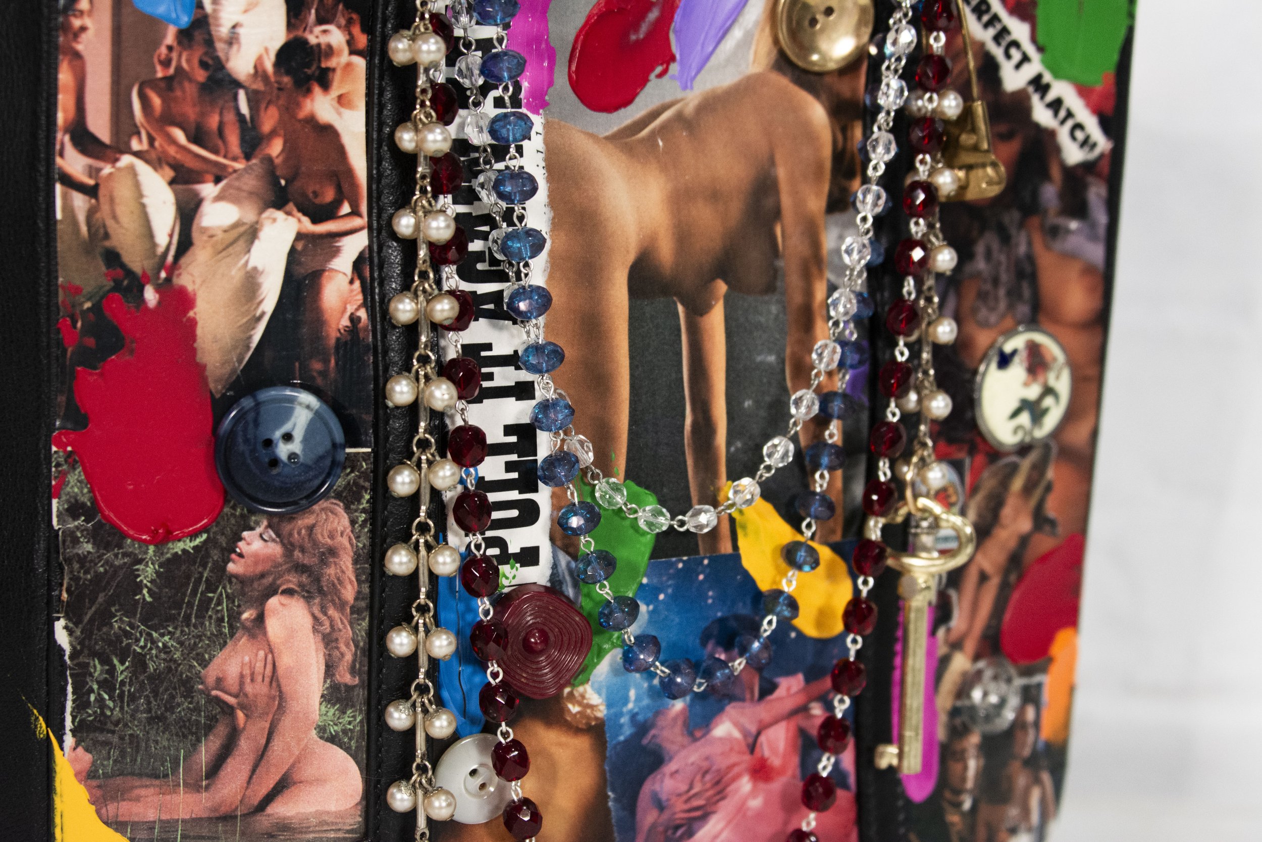 Detail shot of painted, collaged, and sealed Balenciaga black purse with colorful button additions, various charms, beads & hardware being held by it's straps against a white brick wall