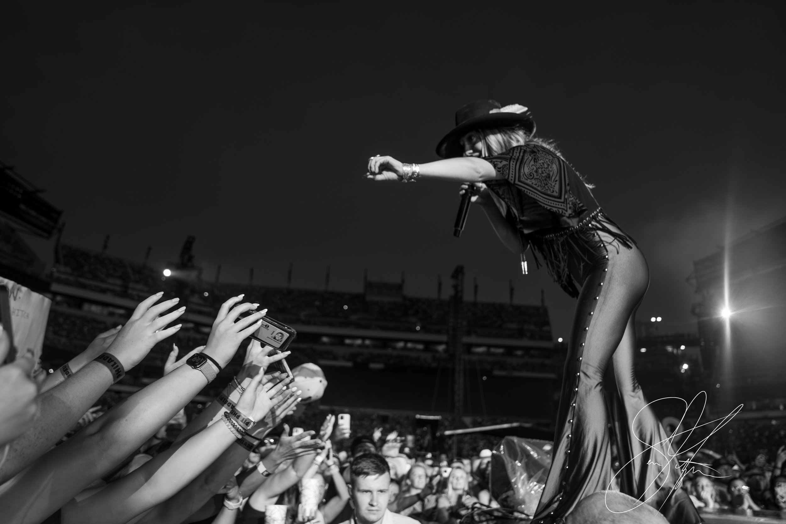 Black and white photo of a musician performing on stage, wearing a hat and holding a microphone, with hands of the audience reaching out and phones raised.