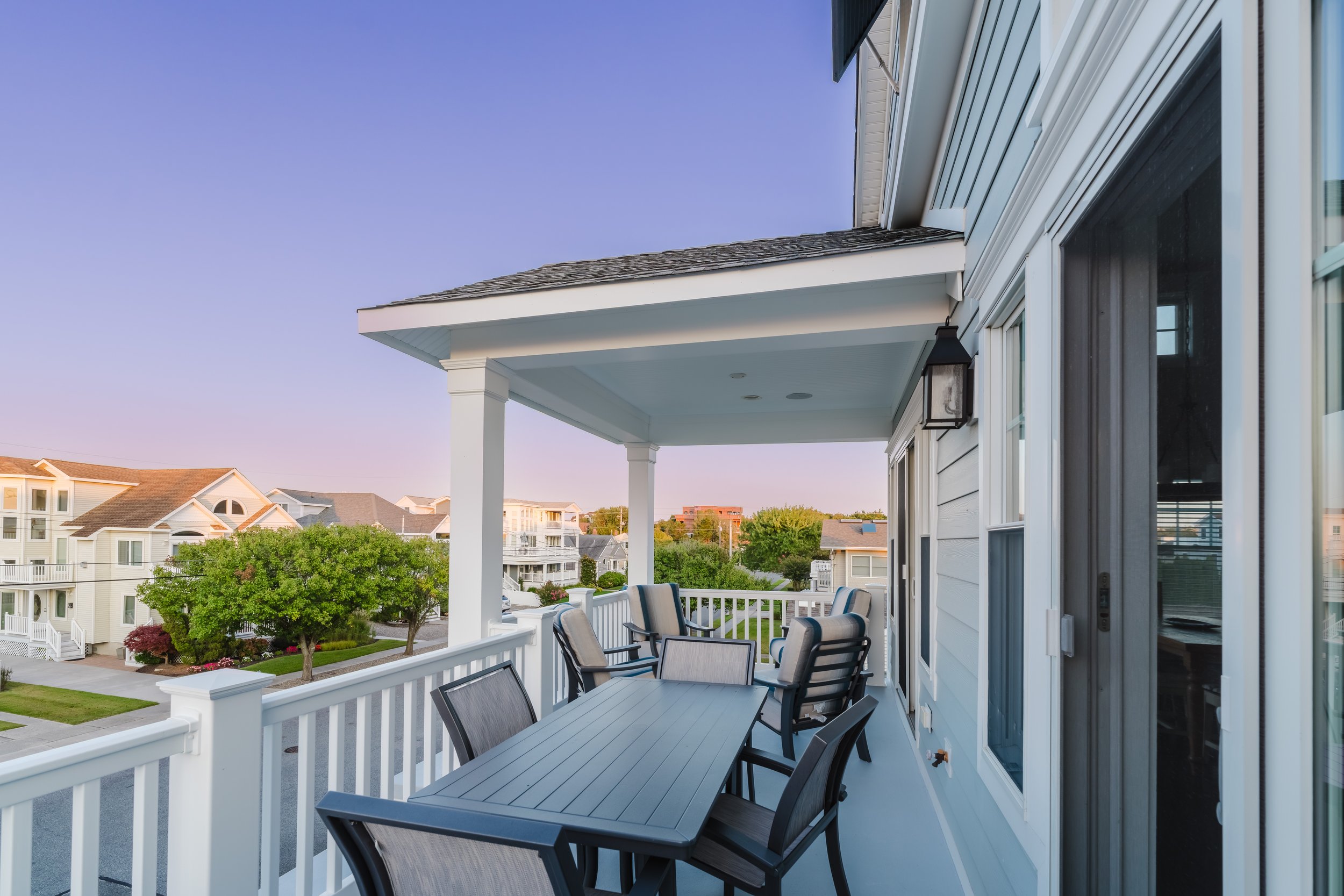 Balcony with table and chairs overlooking houses and trees at sunset.