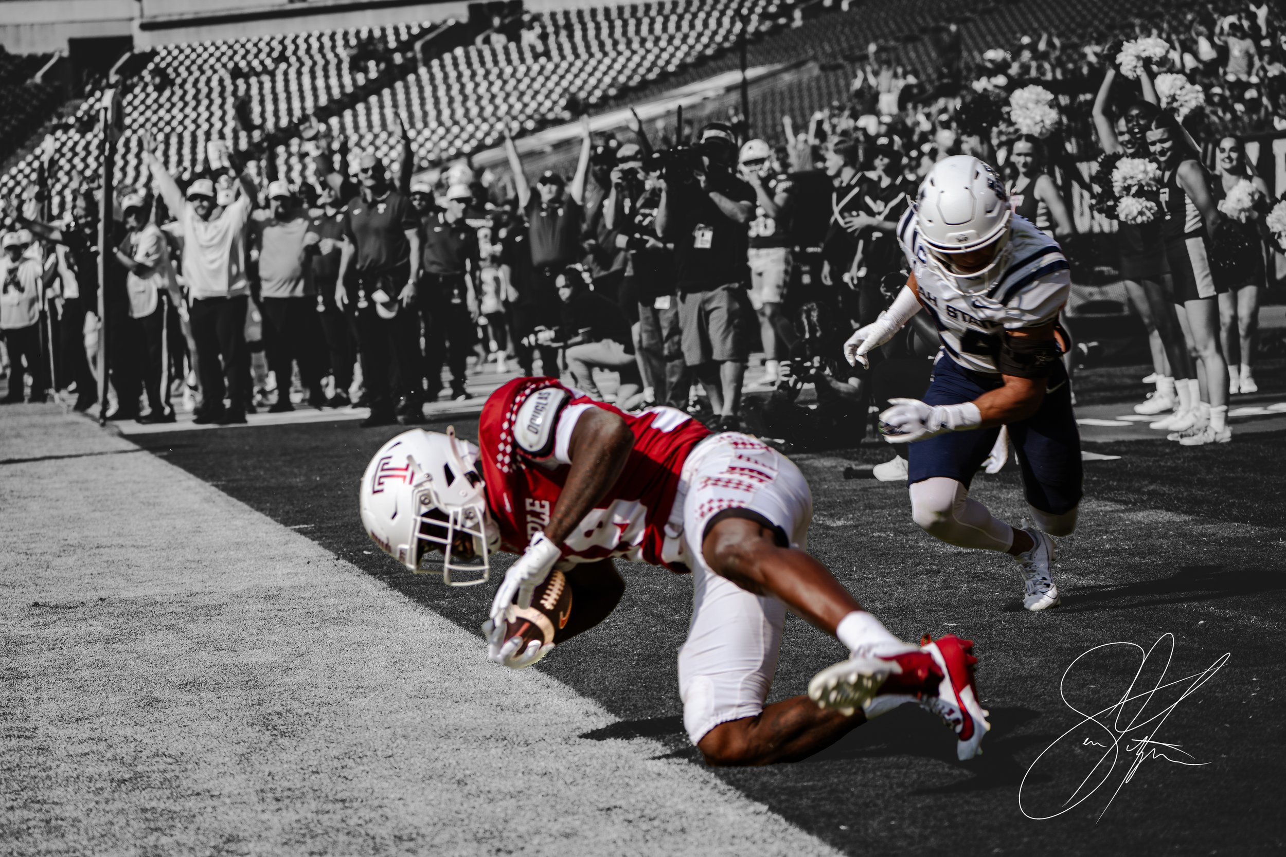 A football player in a red and white uniform dives with the ball towards the end zone during a game, with another player in a blue and white uniform attempting to tackle. The background is monochrome, showing cheering fans and cheerleaders.
