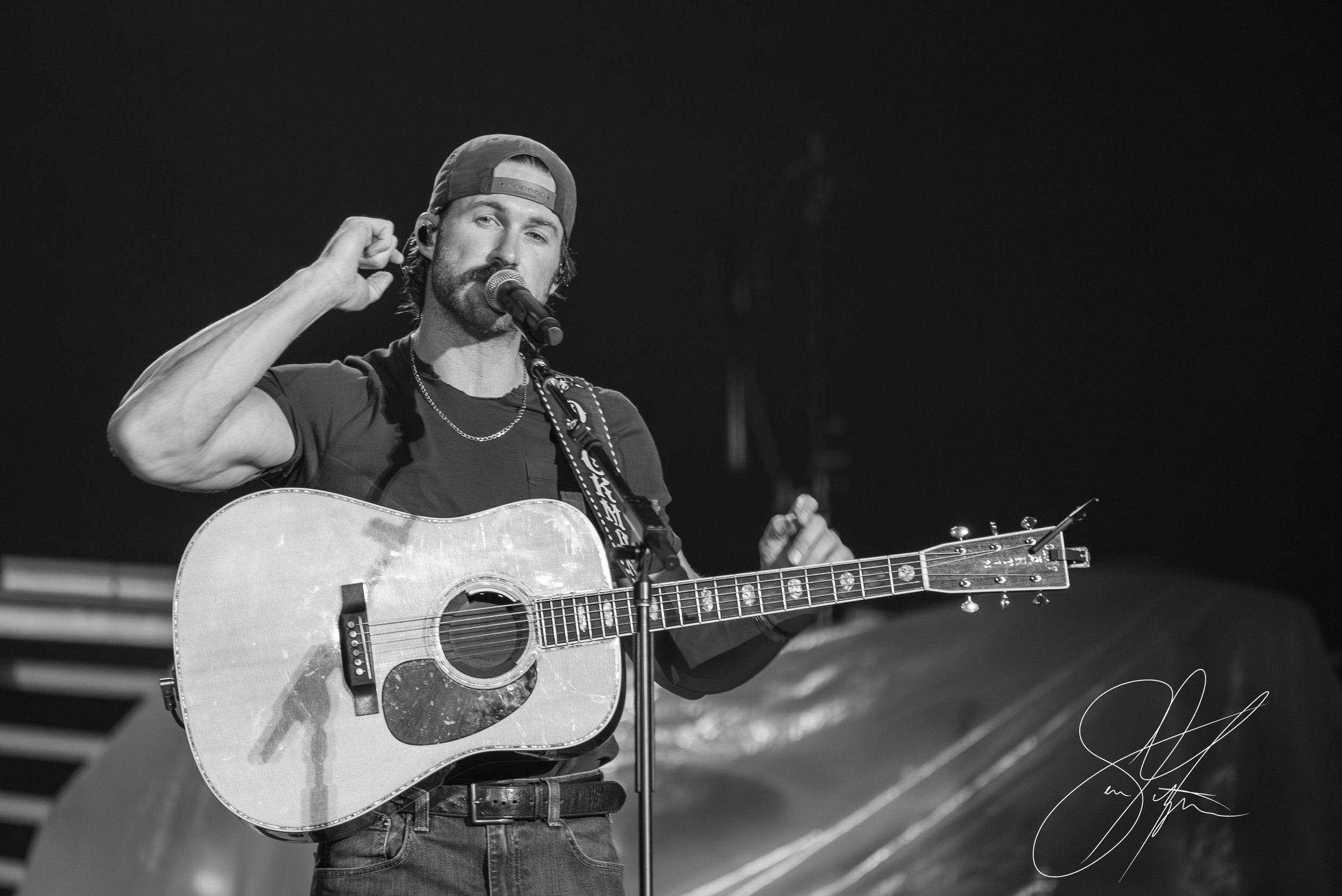 Musician performing on stage with a guitar and microphone, wearing a backwards cap and T-shirt, in black and white.