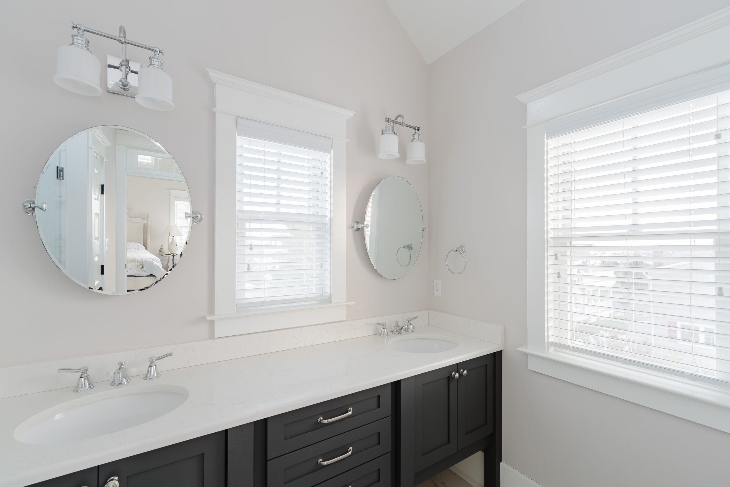 Modern bathroom interior with double sinks, circular mirrors, white countertop, dark cabinetry, and white blinds.