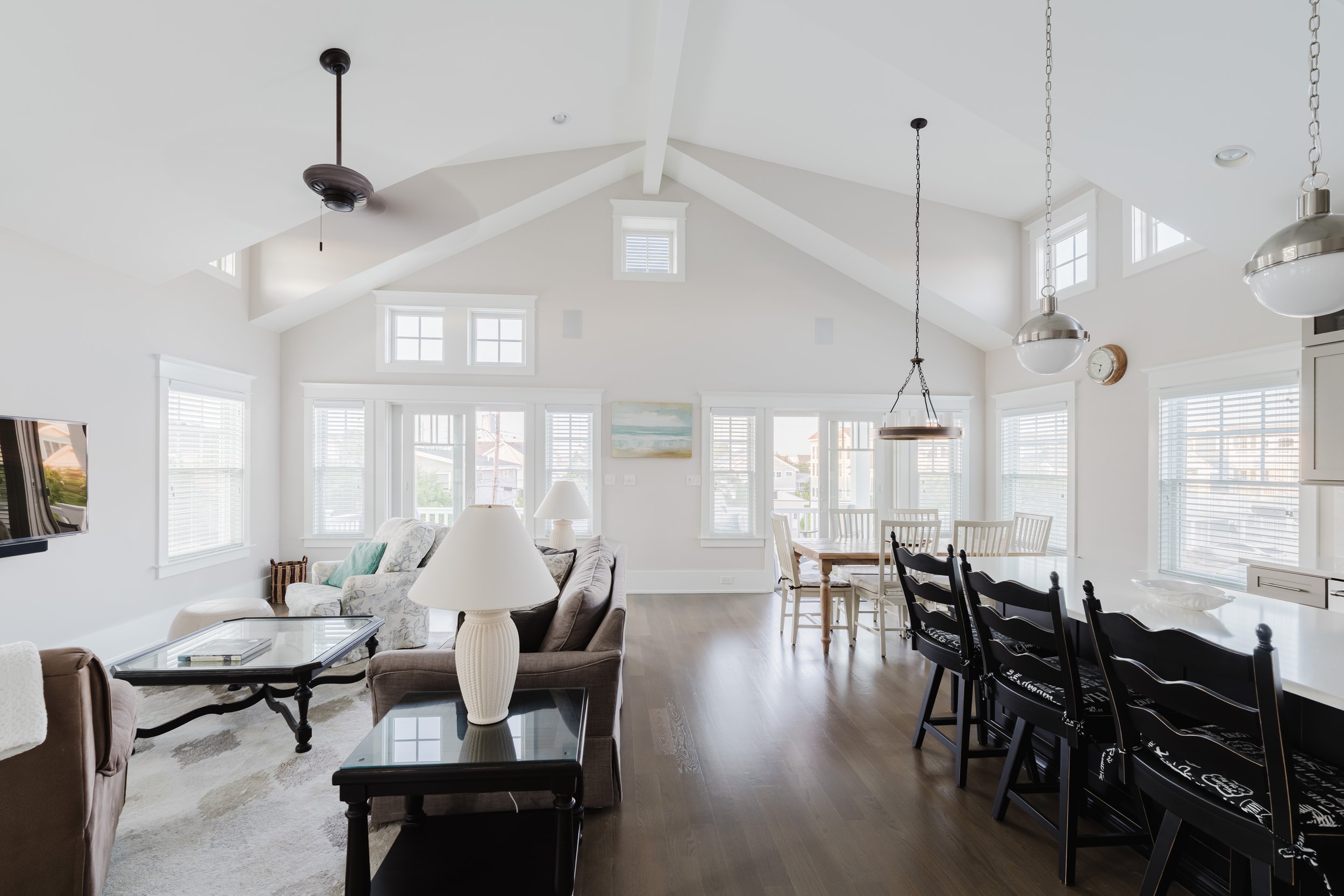 Bright and spacious modern living room with vaulted ceiling, featuring a gray sofa, glass coffee table, ceiling fan, dining area with white chairs, large windows, and two pendant lights.