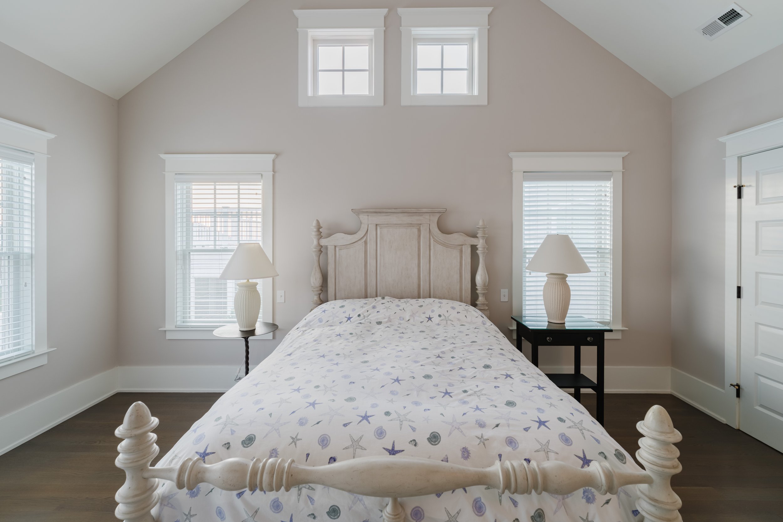 Bedroom with wooden bed, sea-themed bedding, two bedside tables with lamps, and windows with blinds.