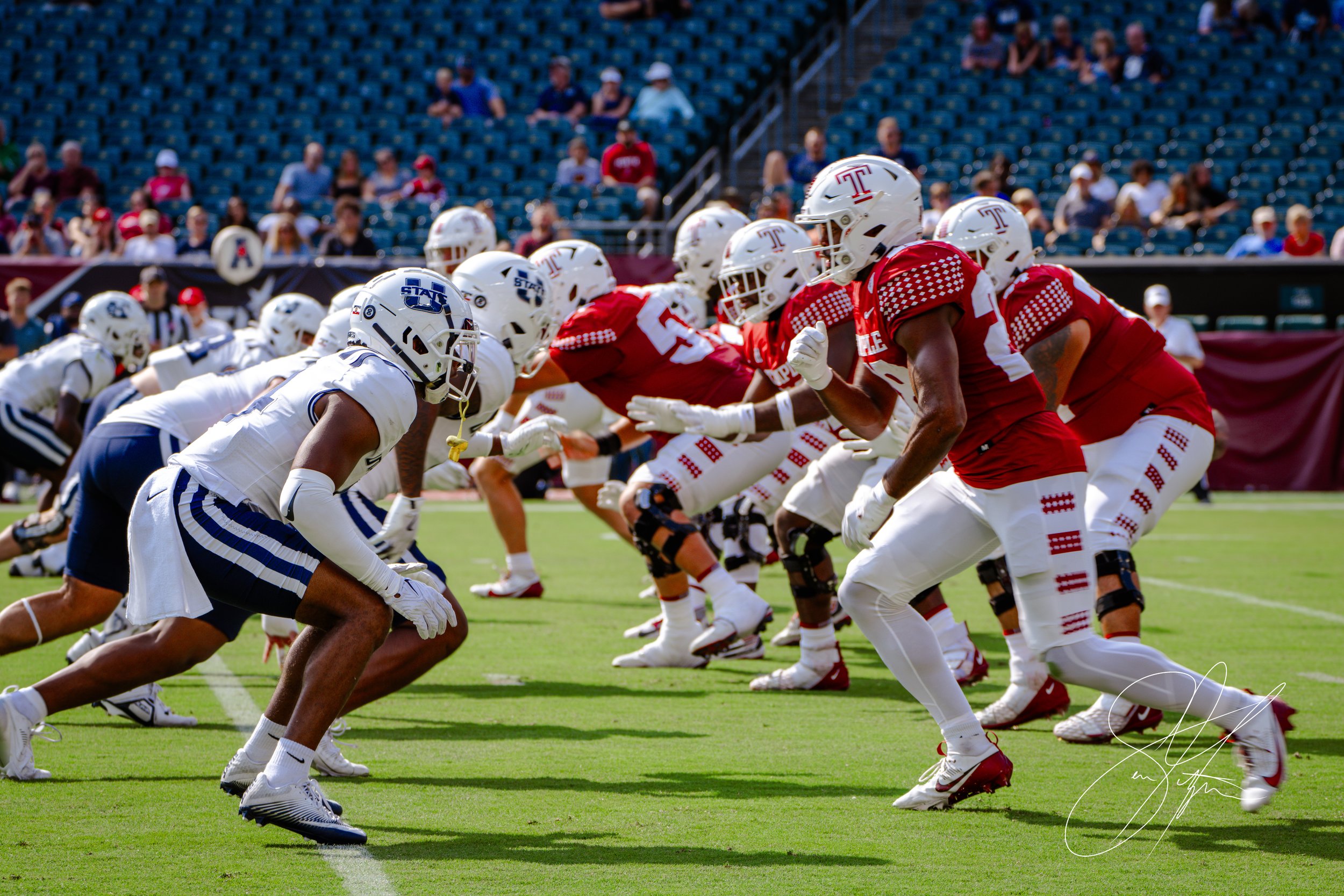 Two college football teams facing off at the line of scrimmage during a game, with one team in red and white uniforms and the other in white and blue, on a field with spectators in the background.