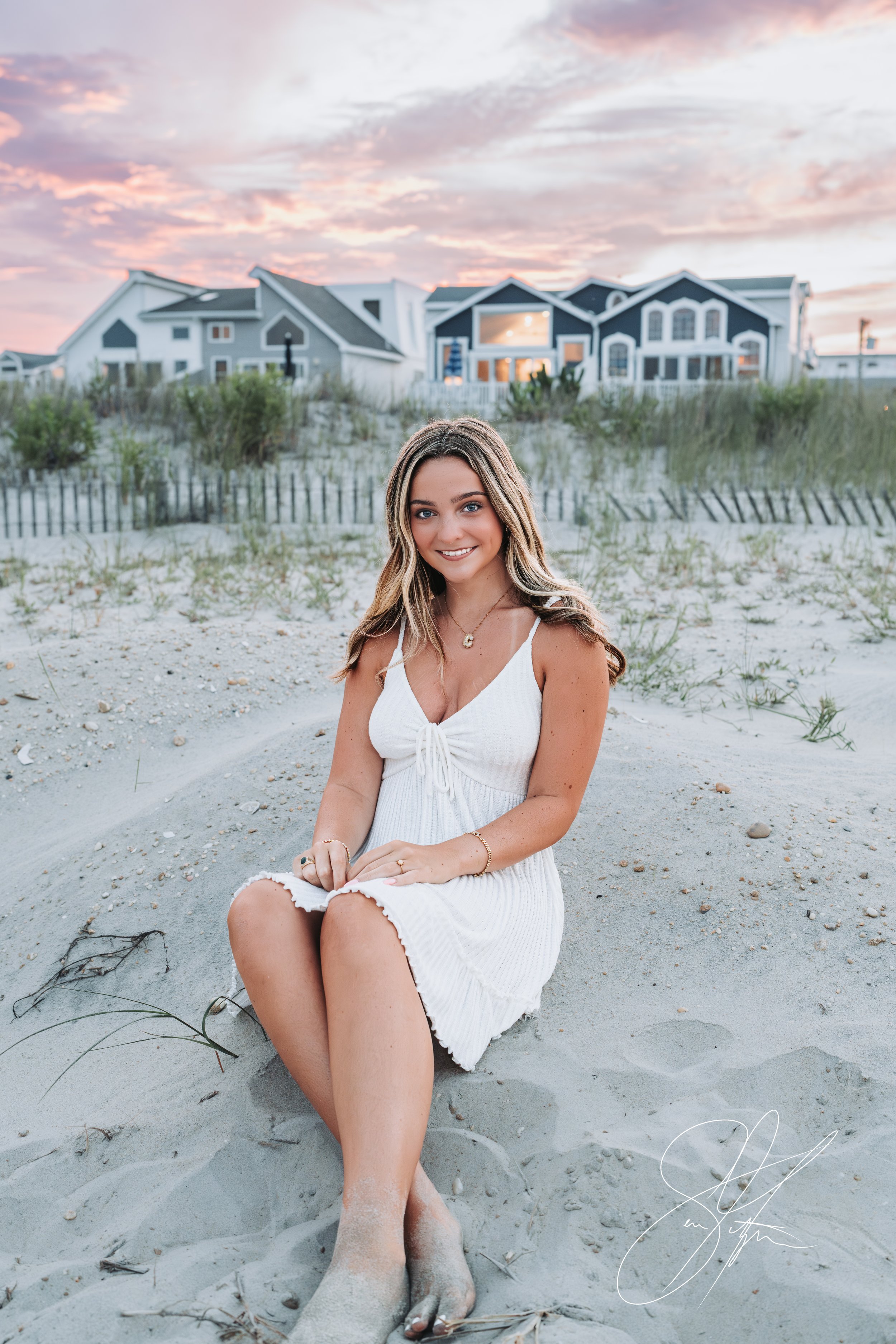 Woman in white dress sitting on sandy beach with grass, houses in the background, and a colorful sunset sky.