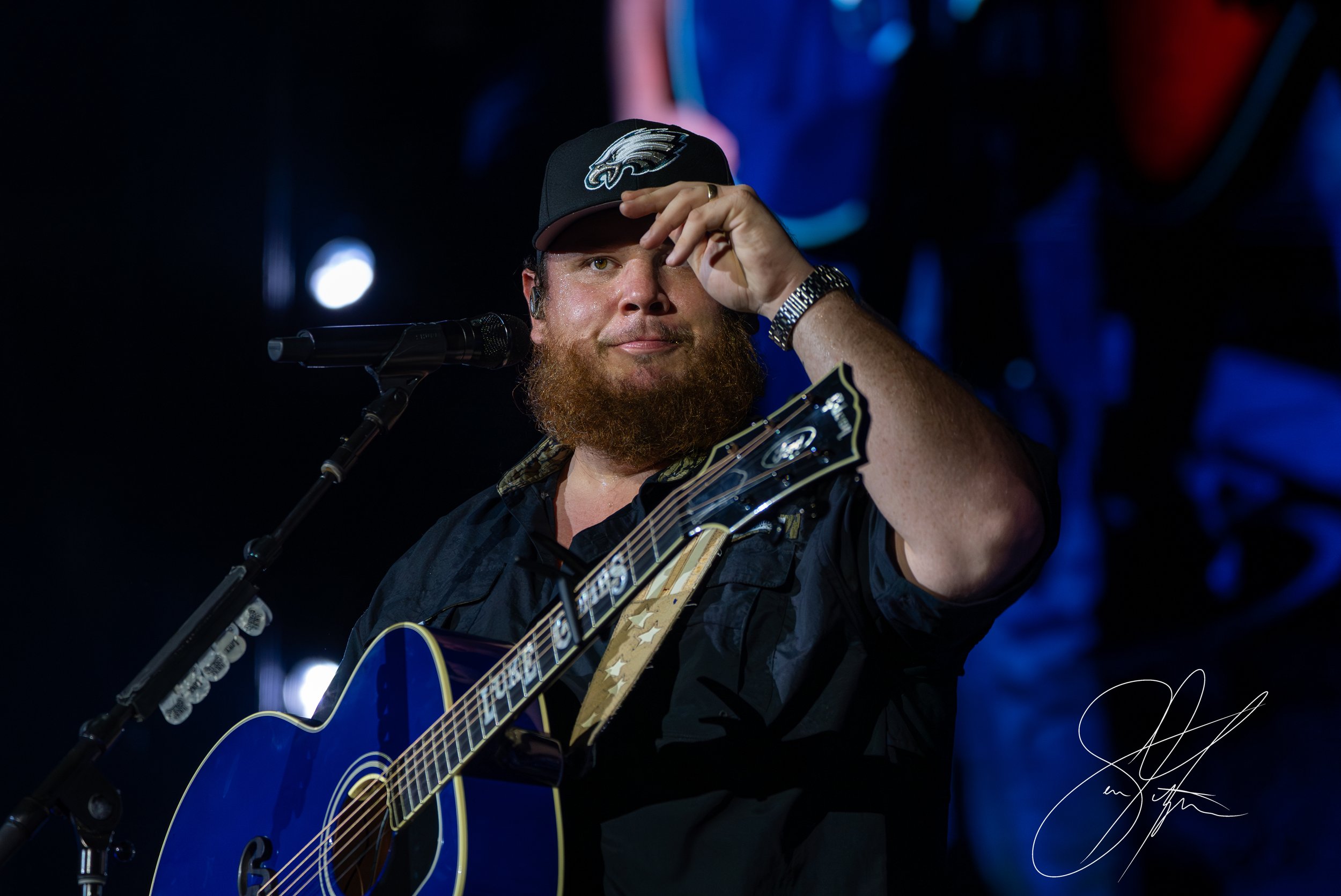 Bearded man wearing a hat and holding a blue guitar on stage with a microphone.