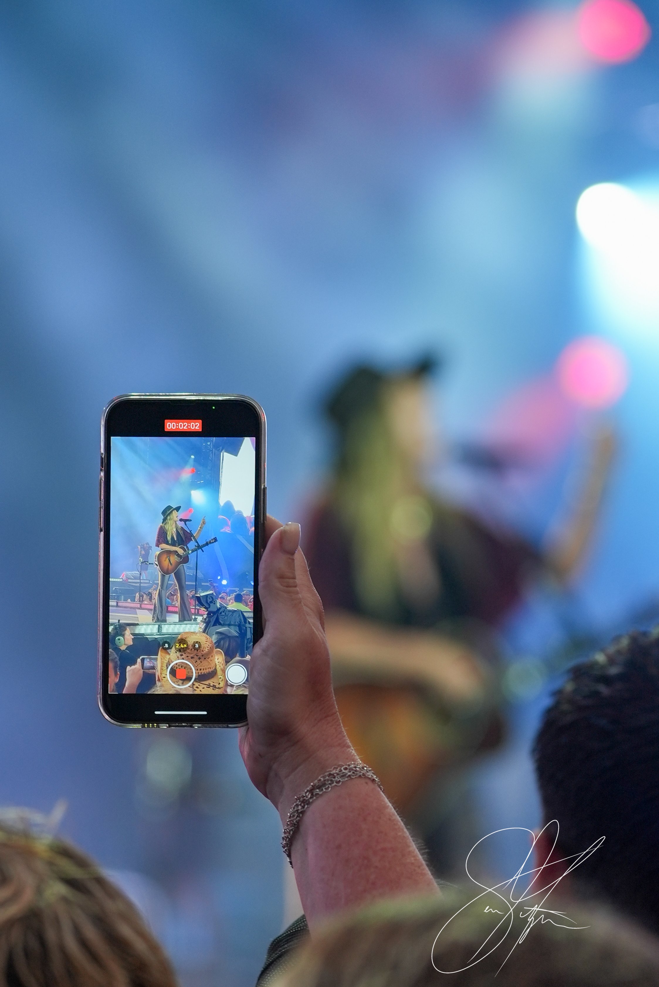 A person holding a smartphone recording a live concert. The phone screen shows a musician on stage with a guitar, wearing a hat. The background is blurred with colorful stage lighting.