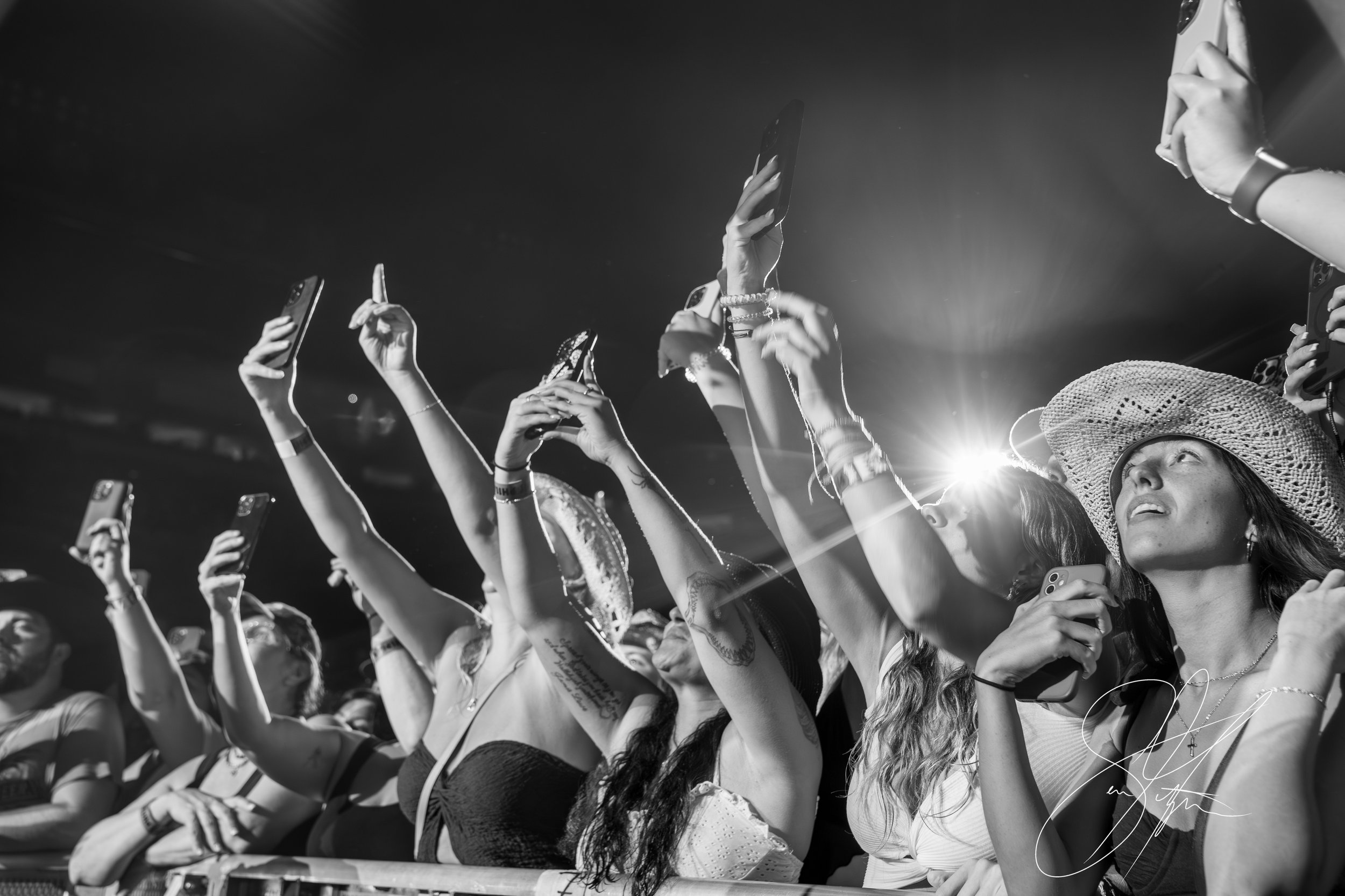 Black and white photo of music concert crowd with hands and phones raised, light from stage illuminating scene, woman in cowboy hat looks on.