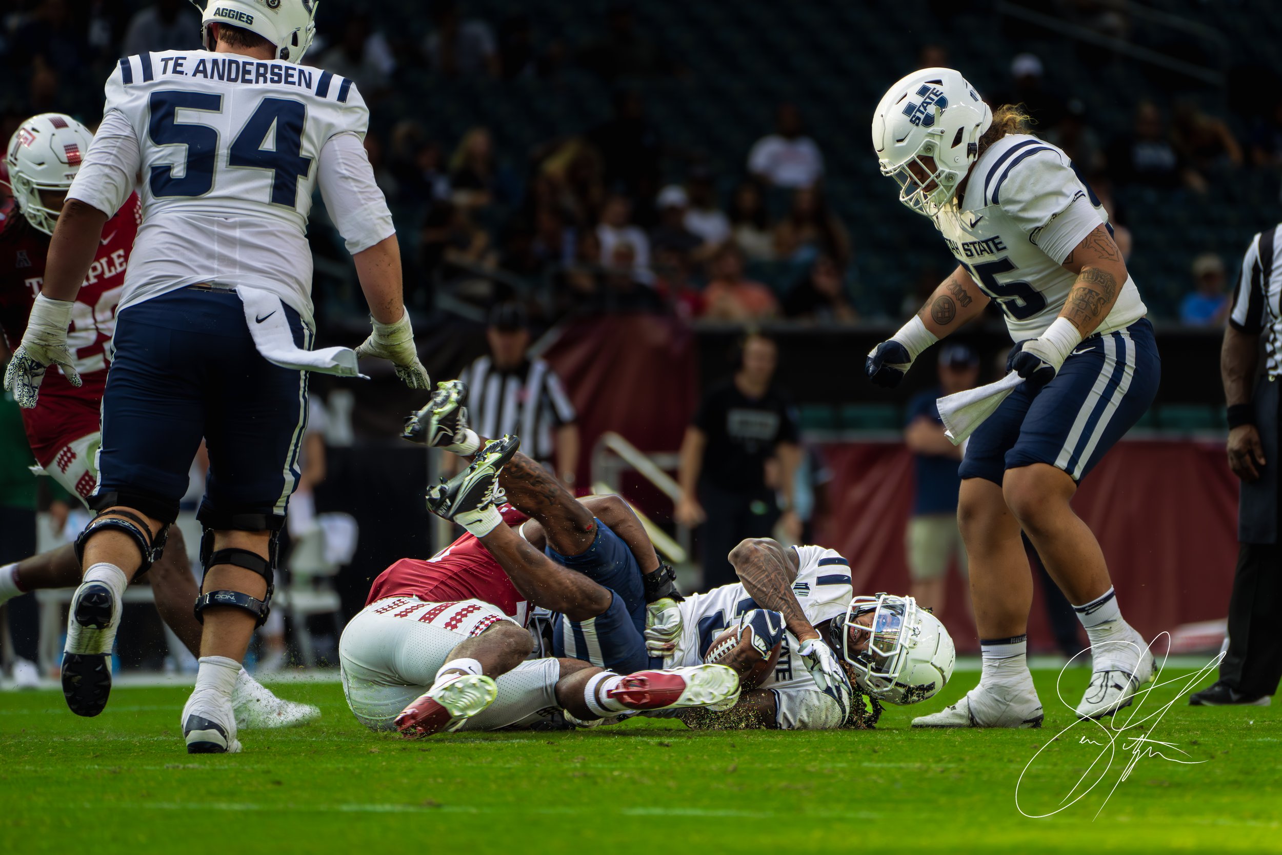 Football players during a game, with one player being tackled. Players are wearing uniforms in blue and white and red and white. An official stands nearby.