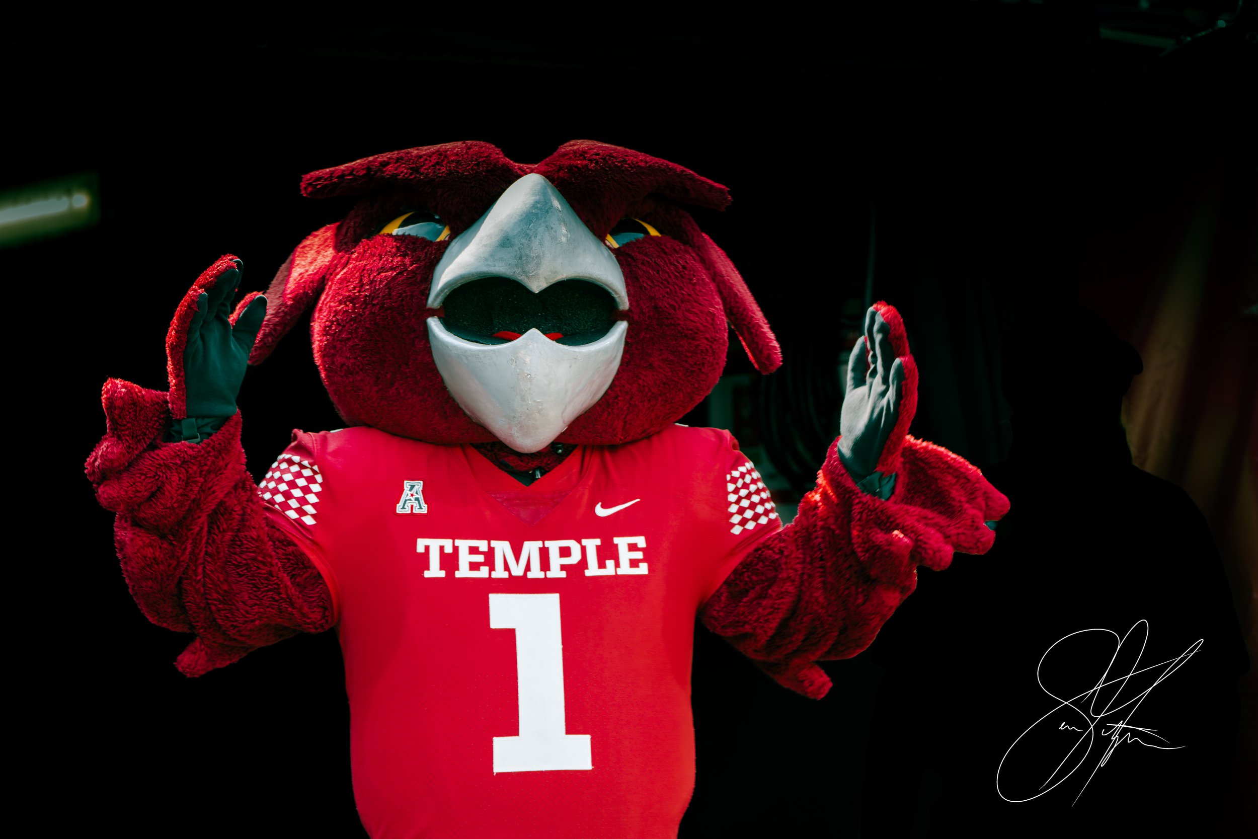 A mascot dressed in a red uniform with the word 'Temple' and the number 1 on the jersey. The mascot has a large beak and is making a gesture with both hands.