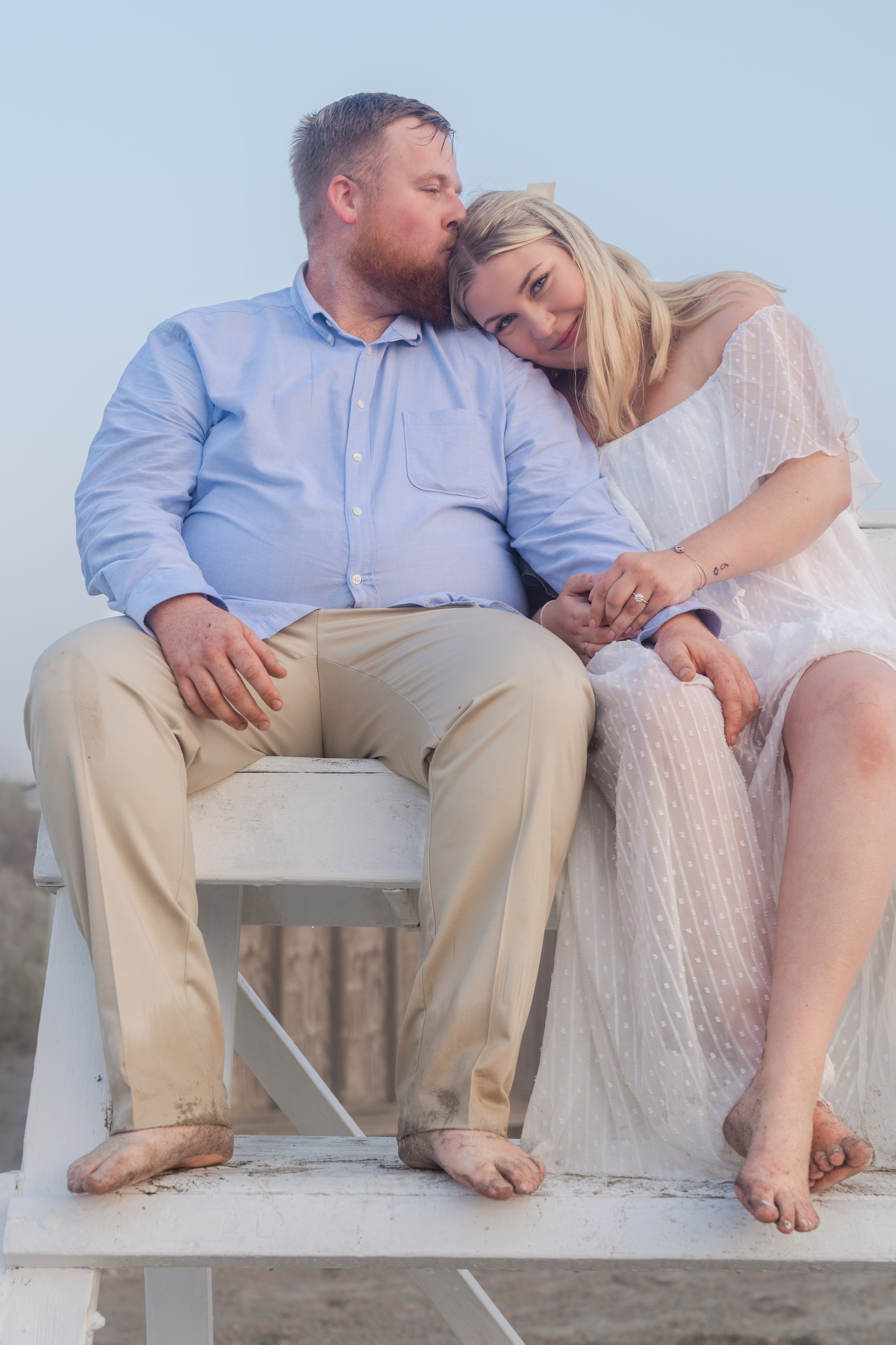 A couple sitting on a wooden bench, with the man kissing the woman's head. The man is wearing a light blue shirt and beige pants, while the woman wears a white dress. Both have barefoot with sandy feet.