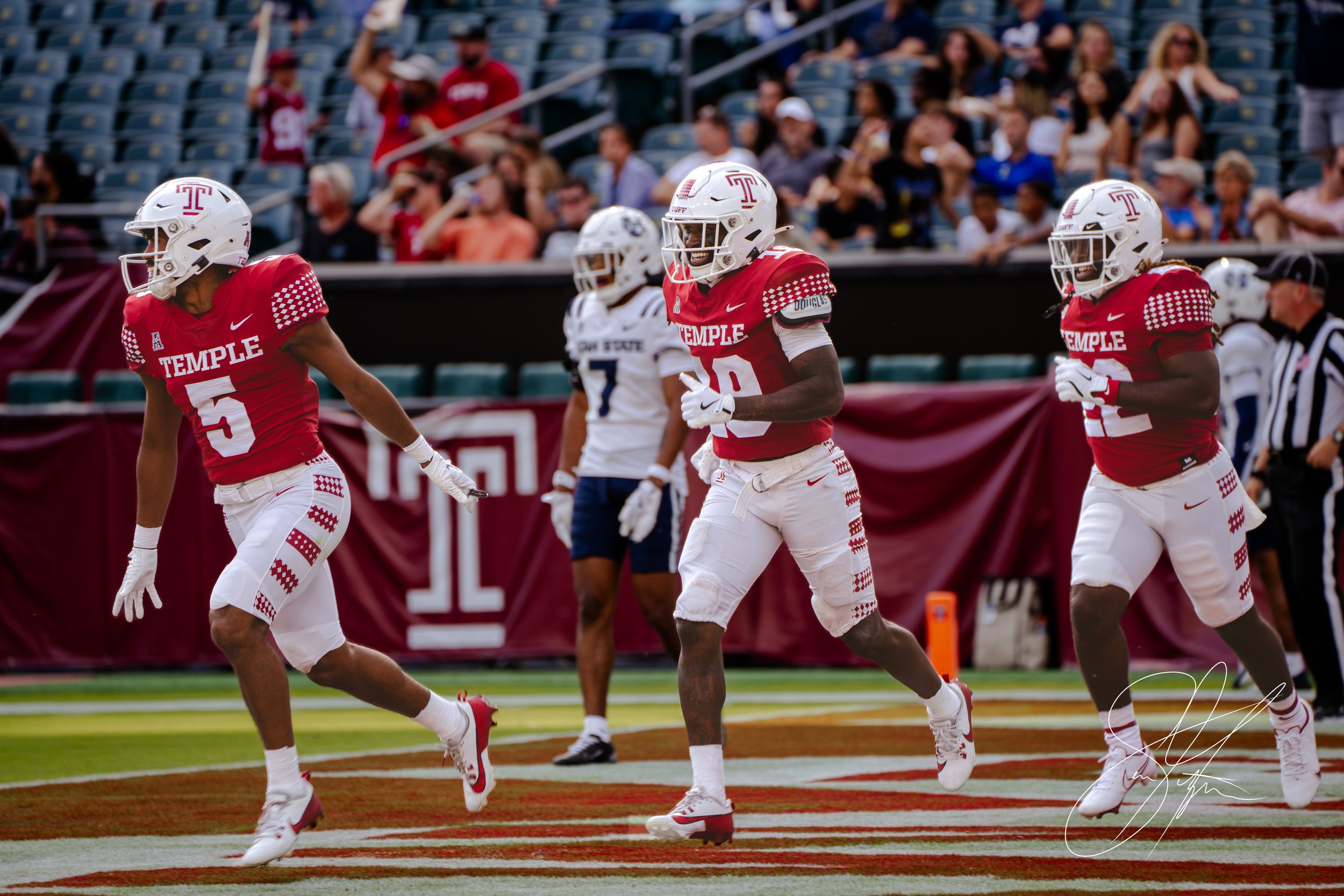 Temple University football players in red uniforms celebrating on the field during a game. A player from the opposing team in white and blue stands nearby. Spectators are seated in the background.