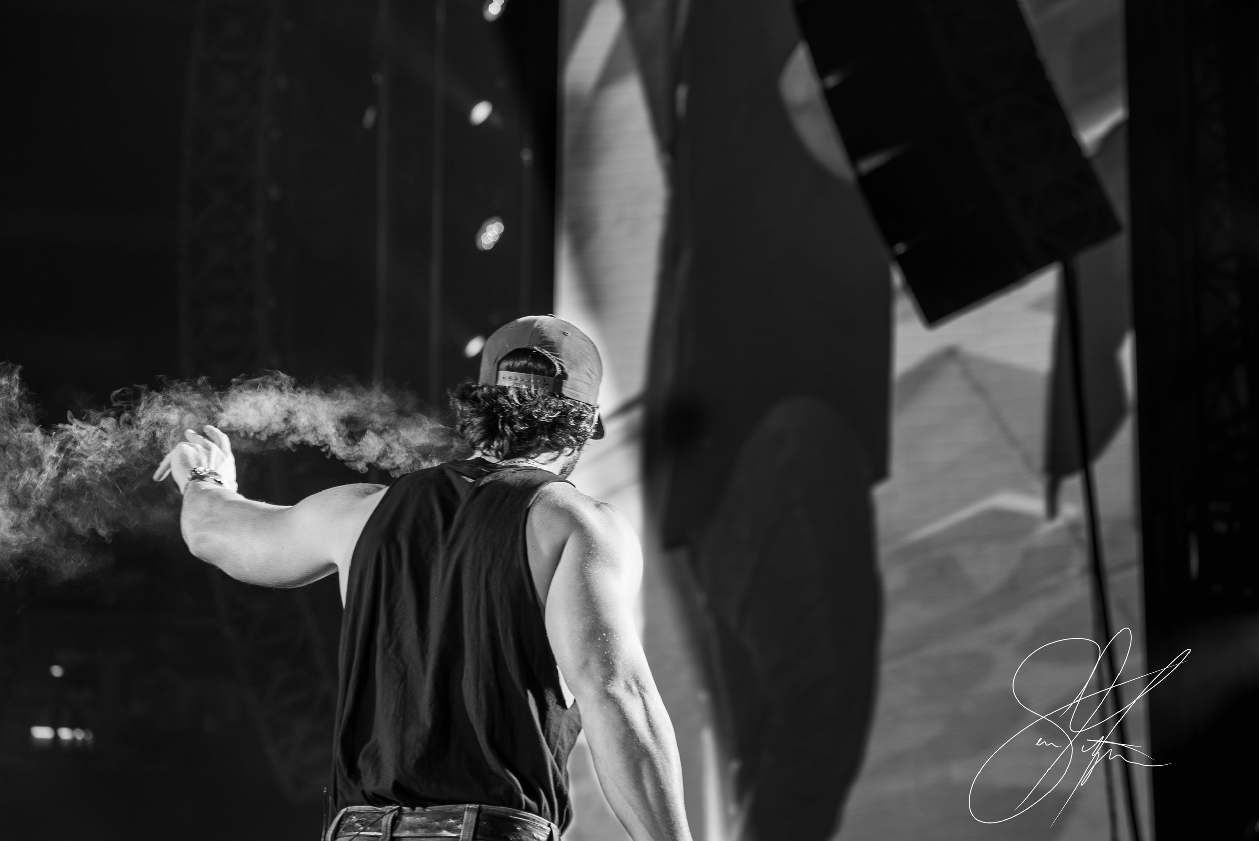 Black and white image of a person with a backward cap and sleeveless shirt on stage, viewed from behind, with a spotlight and shadow on the wall.