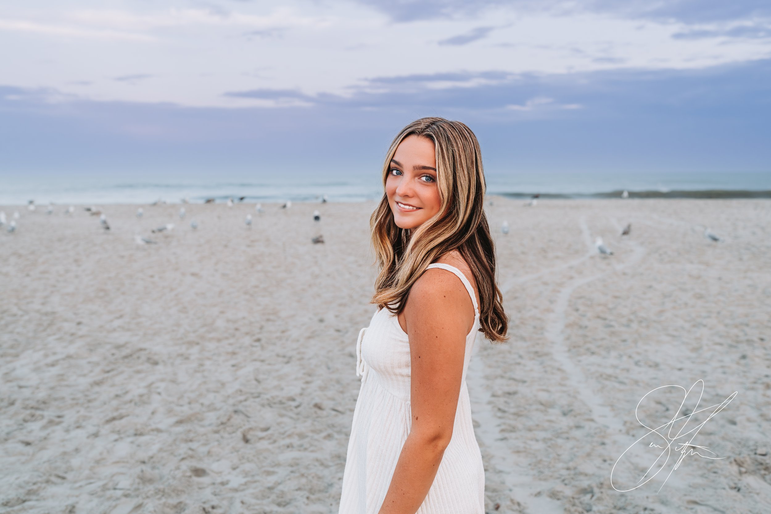 Young woman in white dress smiling on a beach, with seagulls in the background.