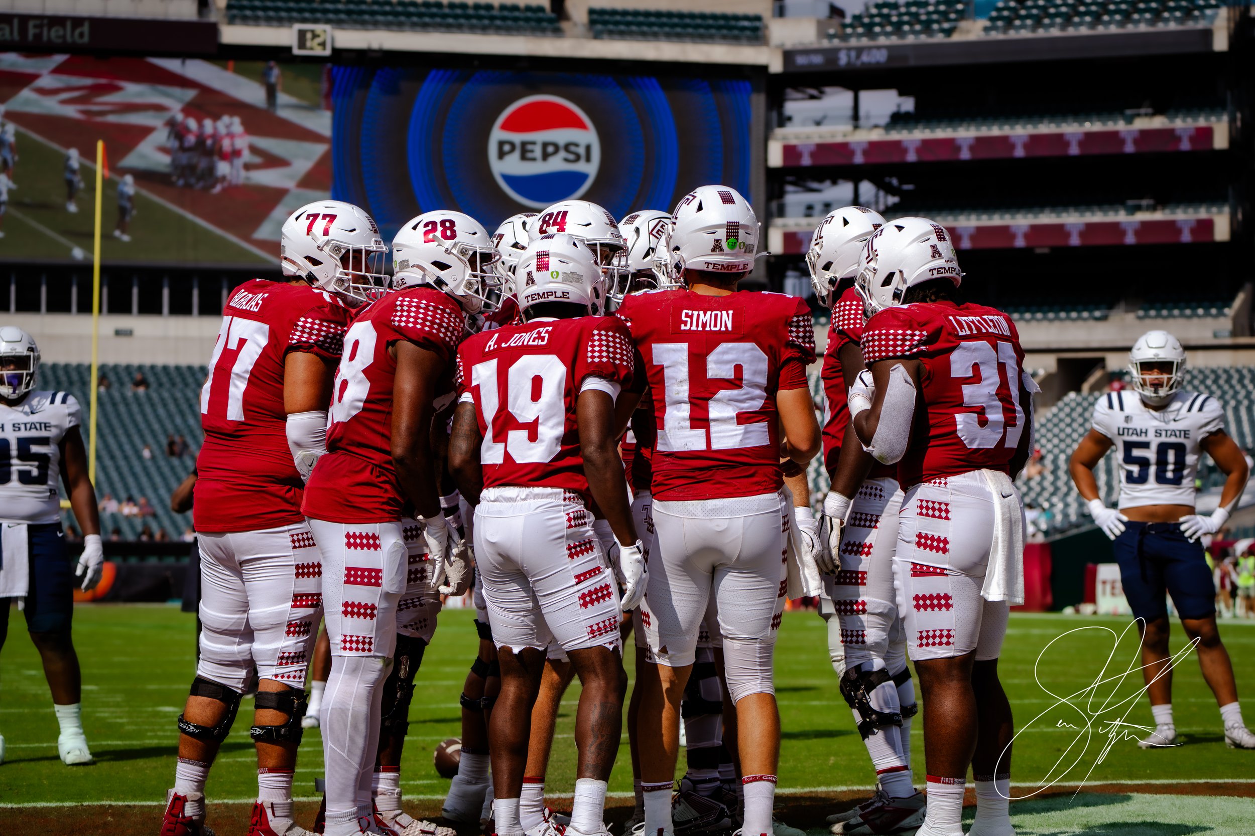 College football players wearing red jerseys in a huddle on the field during a game, with opposing team members from Utah State in white jerseys visible in the background.