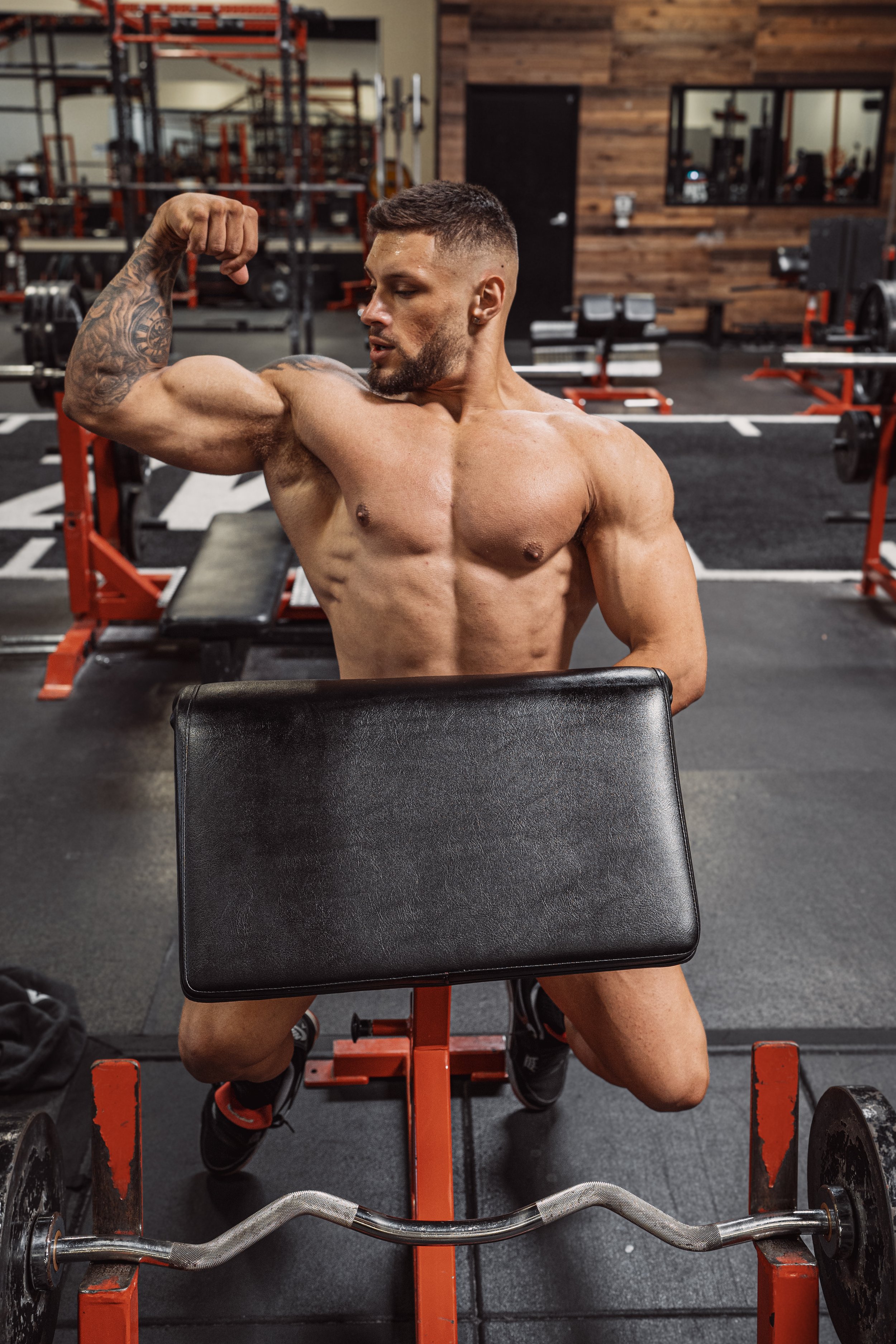 Muscular man flexing biceps in gym, sitting on a preacher curl bench, holding a barbell. Gym equipment visible in the background.