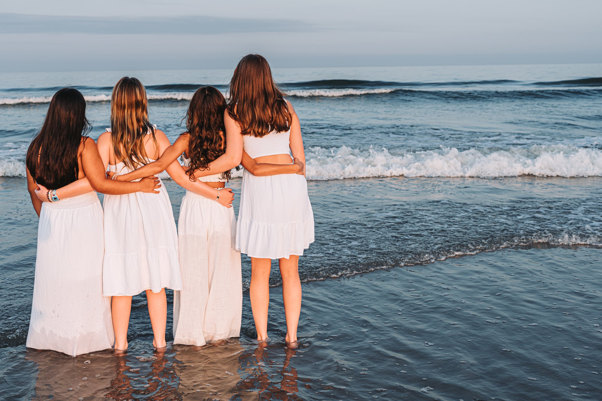 Four women in white dresses standing on a beach facing the ocean, with their arms around each other.