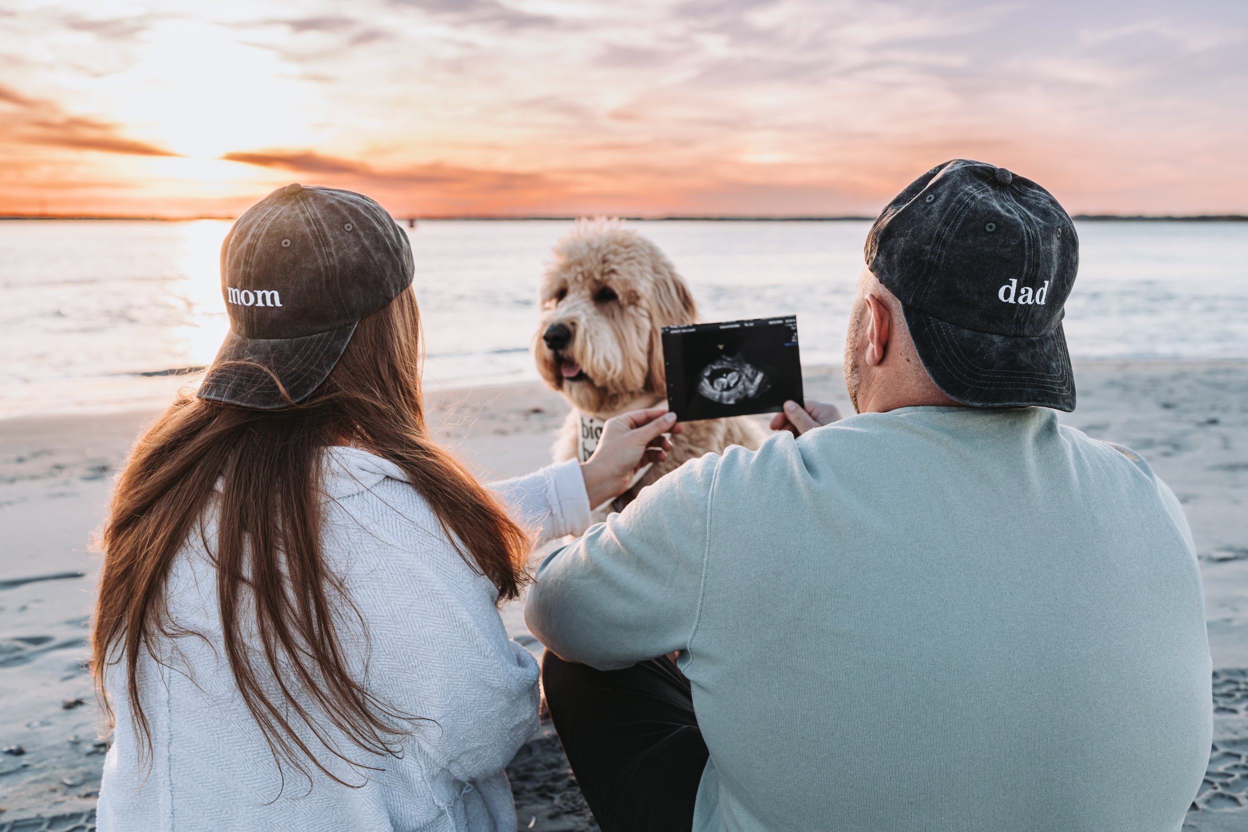 Couple sitting on the beach at sunset, wearing hats labeled 'mom' and 'dad,' holding an ultrasound photo, with a dog nearby.
