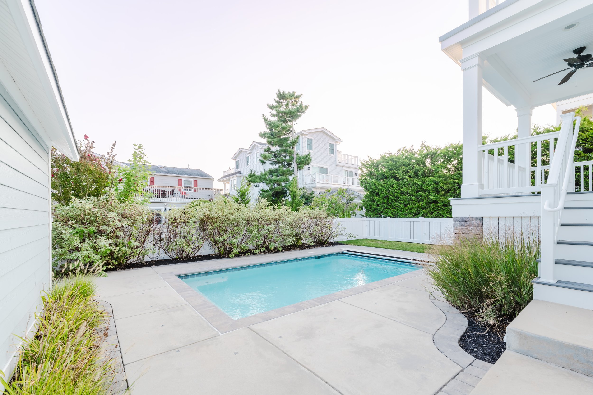 A backyard with a small rectangular swimming pool, surrounded by bushes and a concrete patio area. White fence and neighboring houses in the background, with a porch featuring a ceiling fan and stairs on the right.