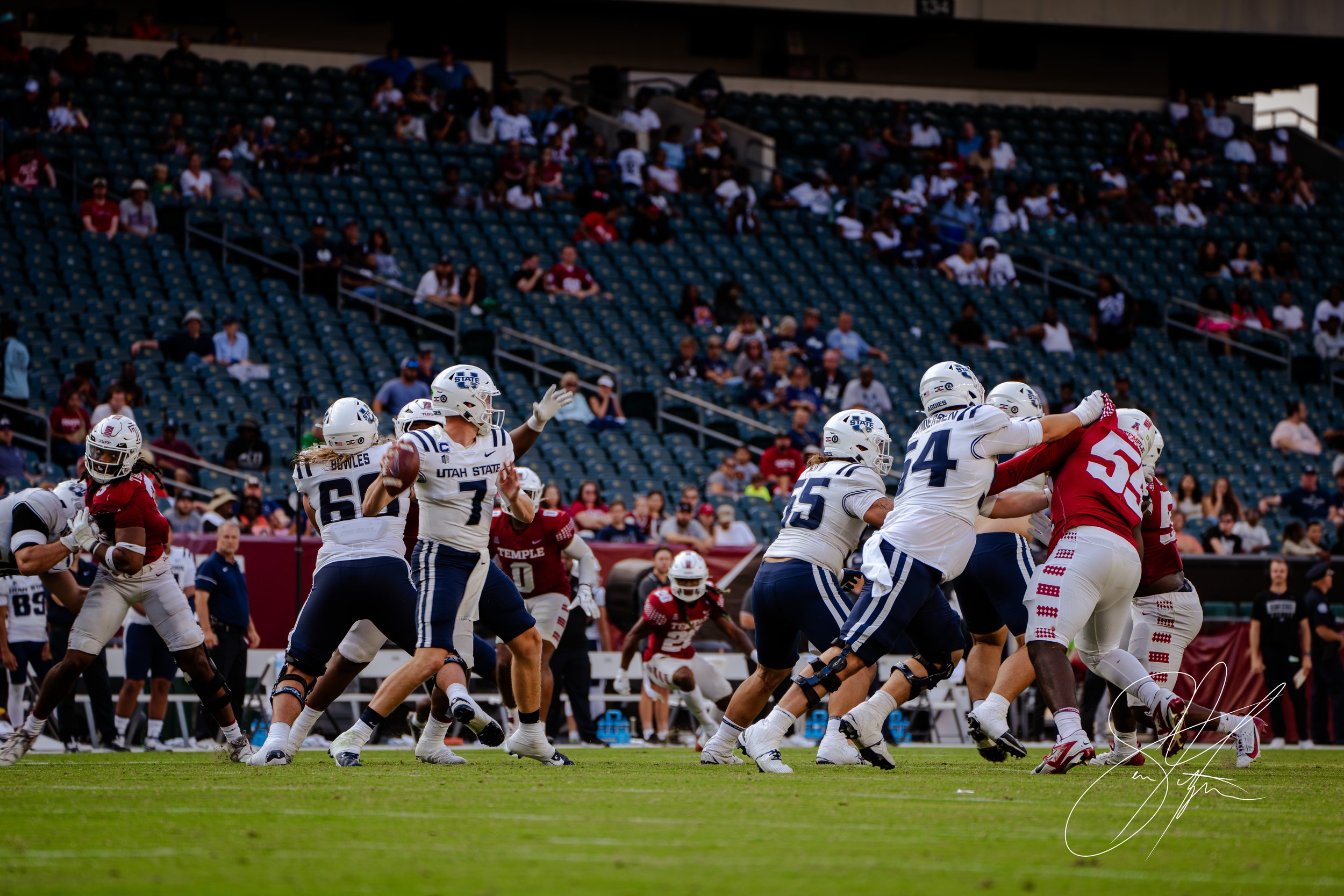 College football game with Utah State players in white uniforms, quarterback preparing to throw, opposing team in red uniforms, stadium in background.