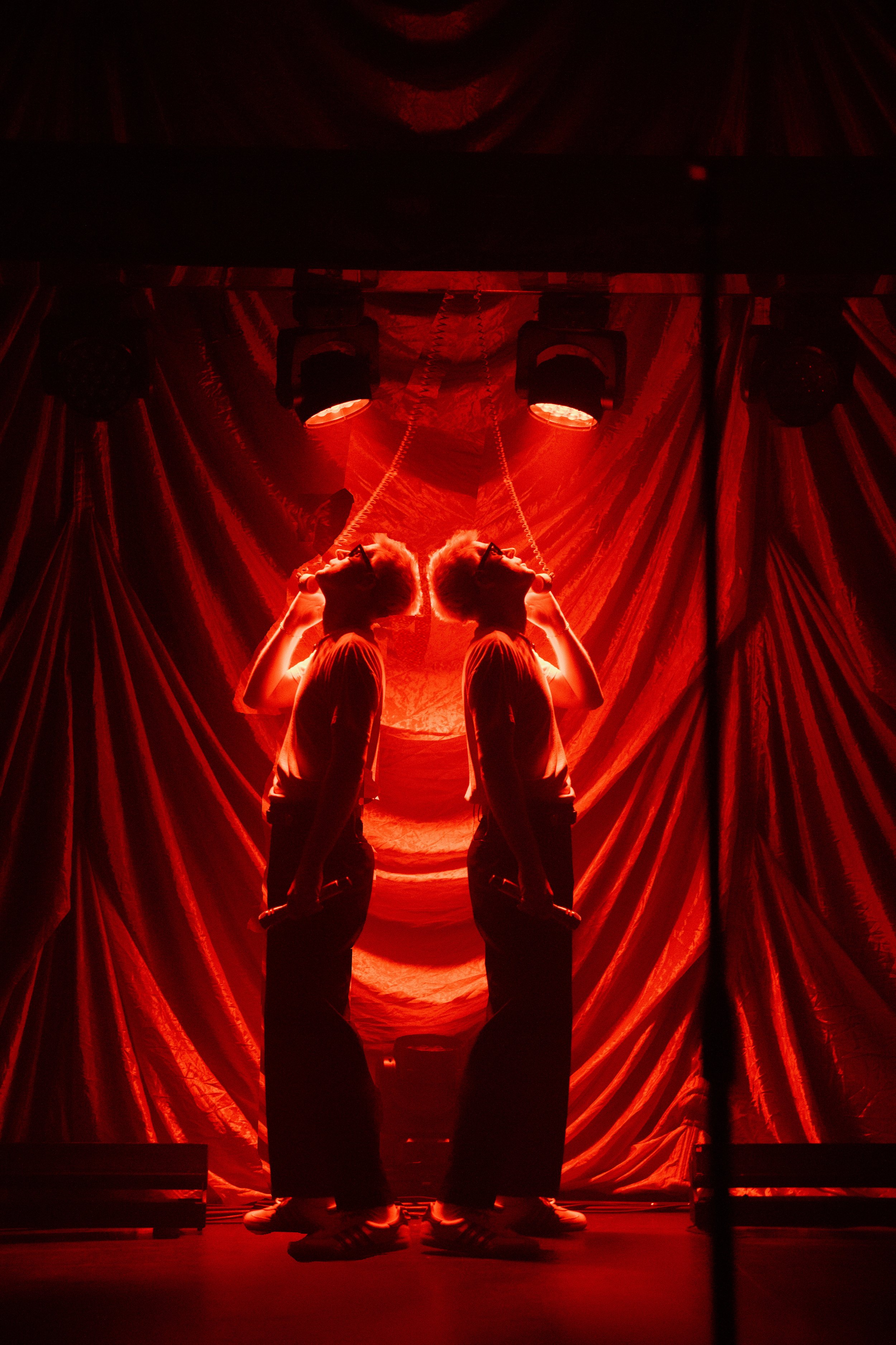 Two mirrored performers under red stage lighting, with draped fabric background, each holding a microphone.