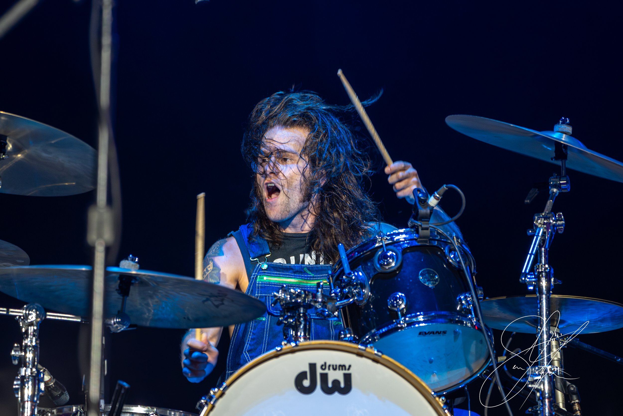 Drummer energetically playing a drum set on stage, holding drumsticks with long hair covering part of their face, wearing a sleeveless shirt and overalls.