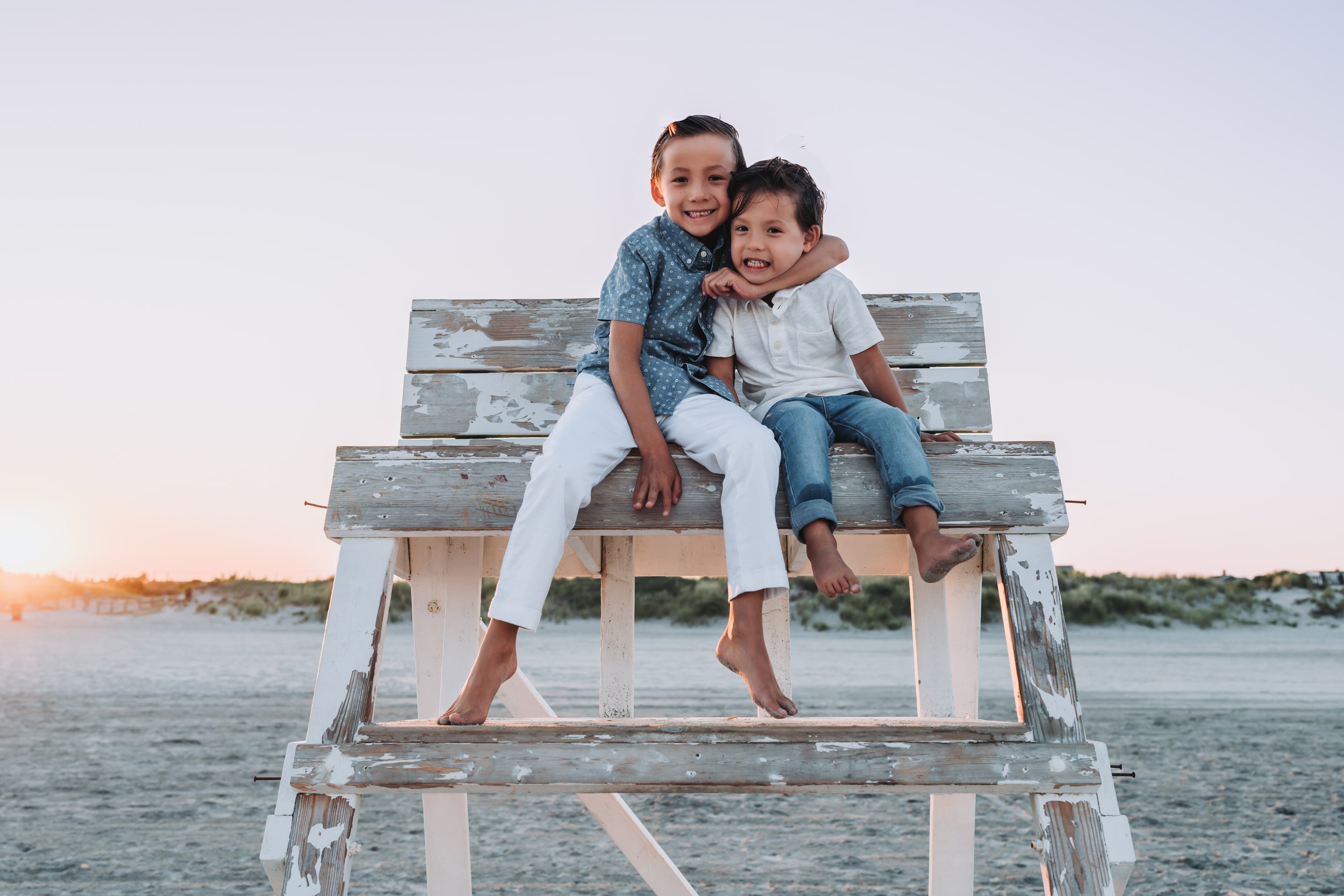 Two children smiling and sitting on a weathered wooden lifeguard chair at the beach during sunset.