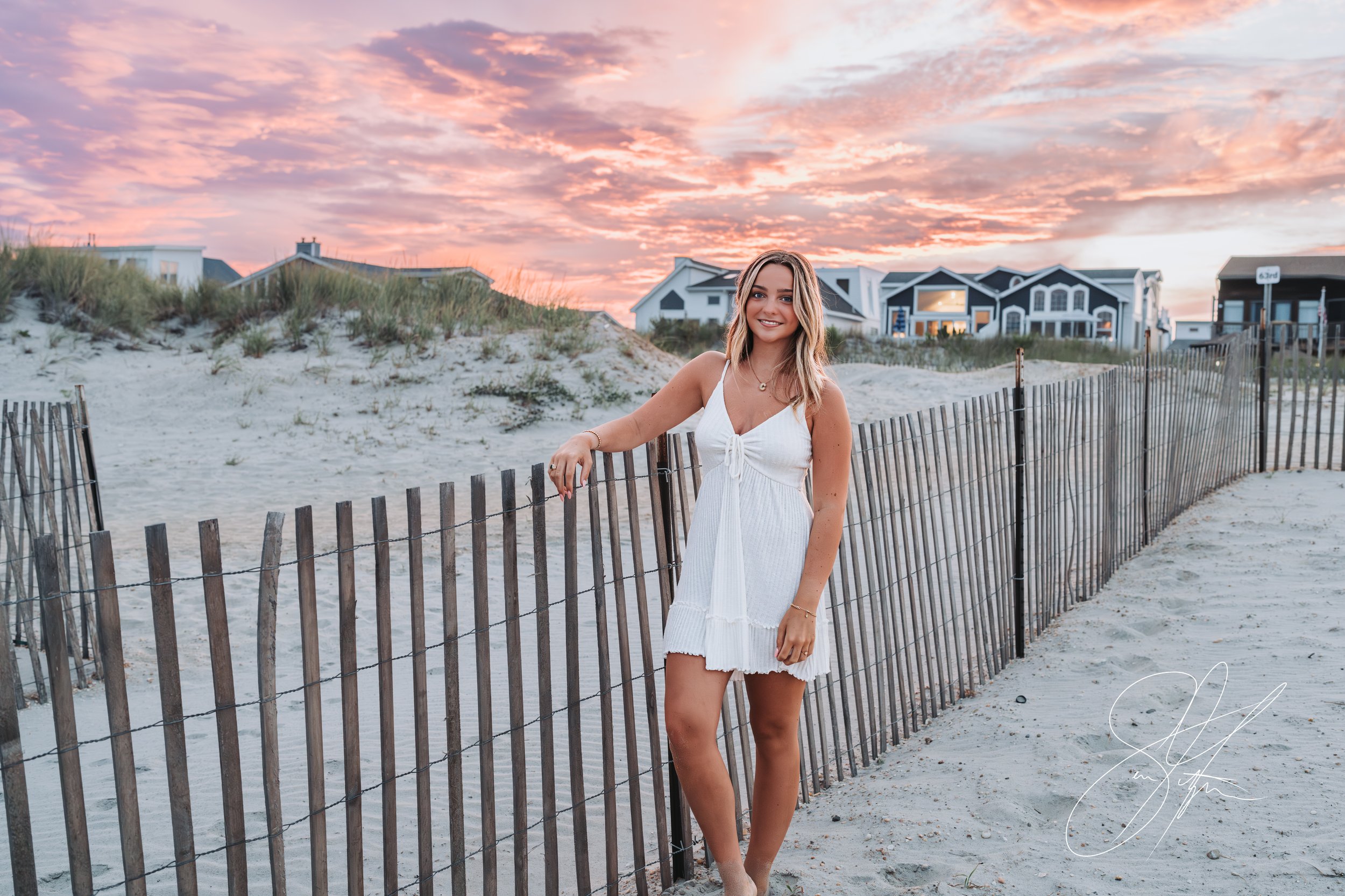 Woman in white dress standing by a wooden fence on a sandy beach at sunset with houses in the background.