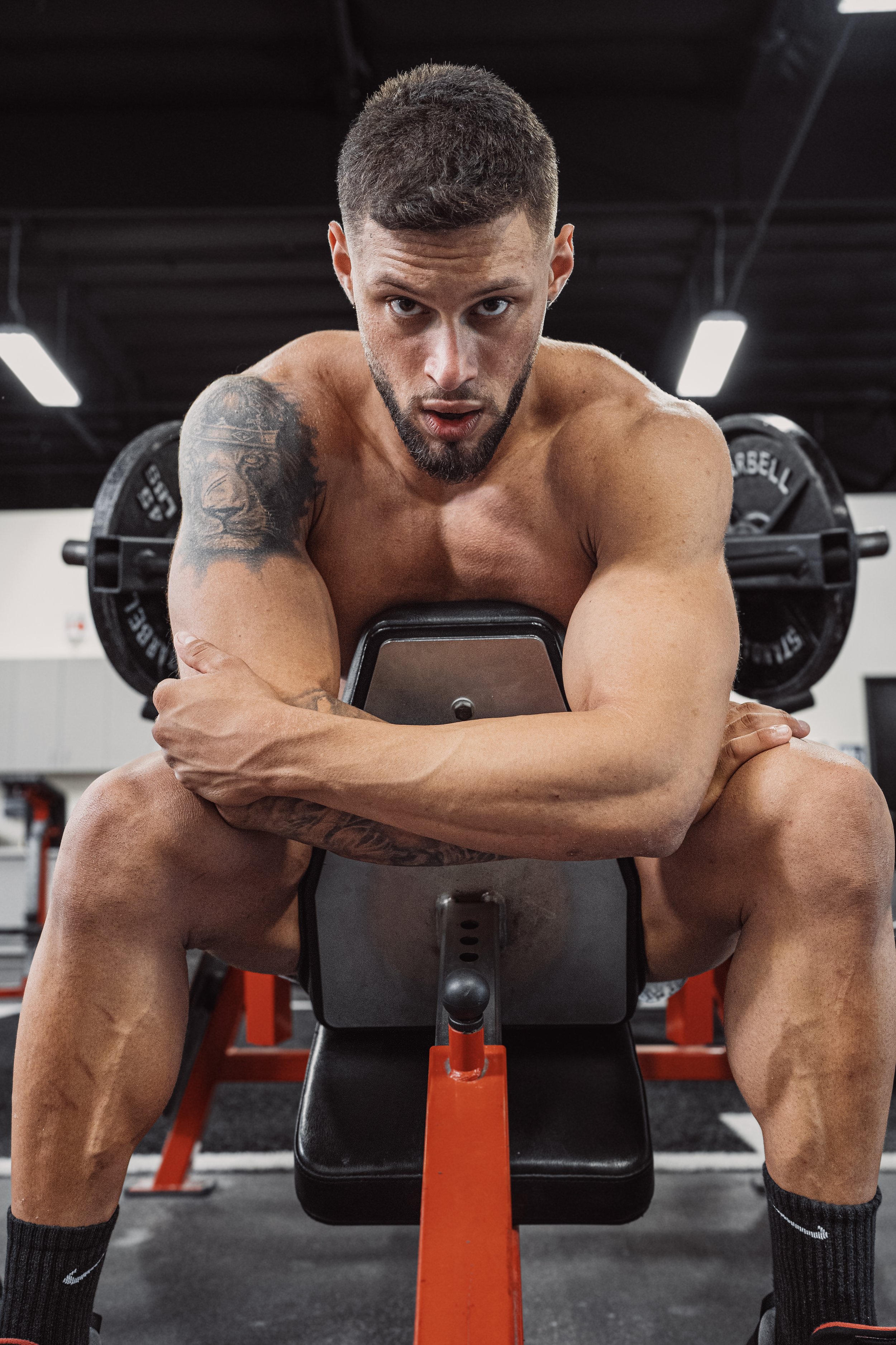 Man with beard and tattoos sitting on gym equipment, looking serious.