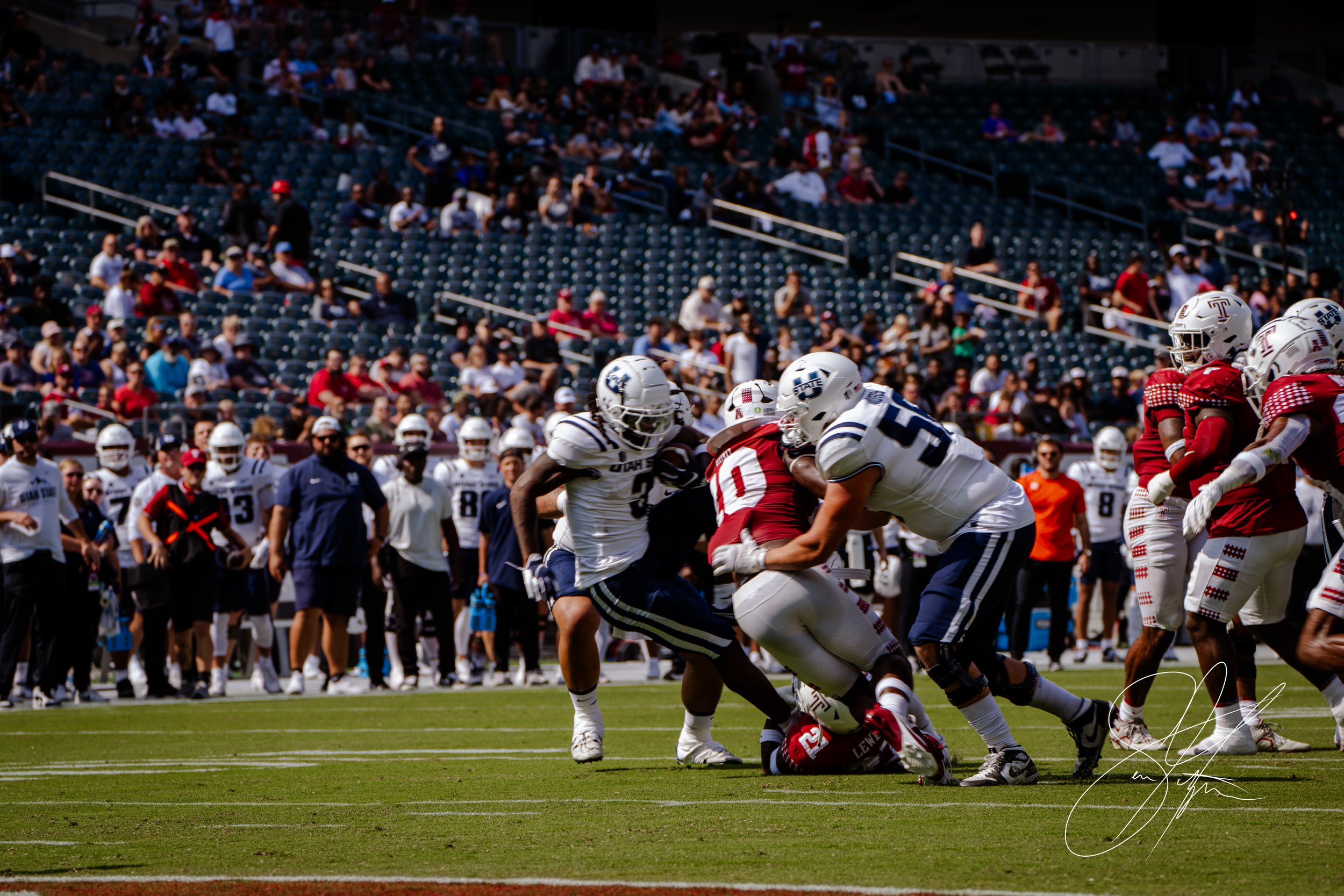 American football players from two teams, in white and red uniforms, in action during a game. One player in white is tackling a player in red. A crowd and team members are visible in the background.