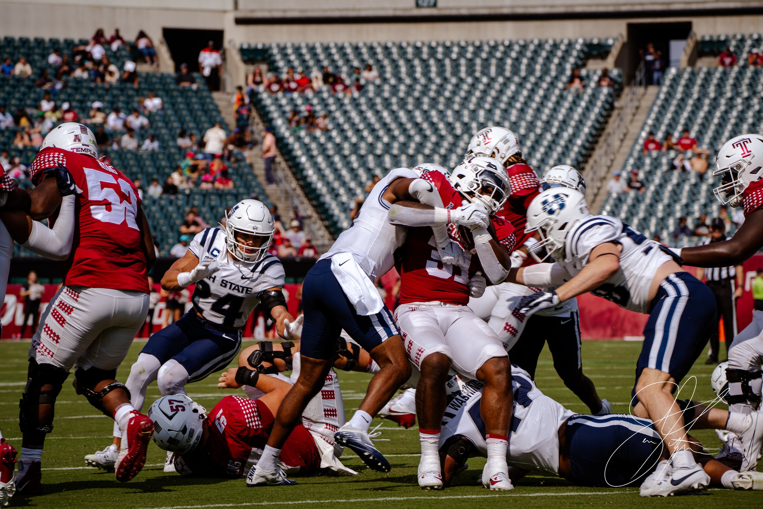 College football game action with players in red jerseys from Temple and blue jerseys from Utah State in a tackle. Stadium seats visible in the background.