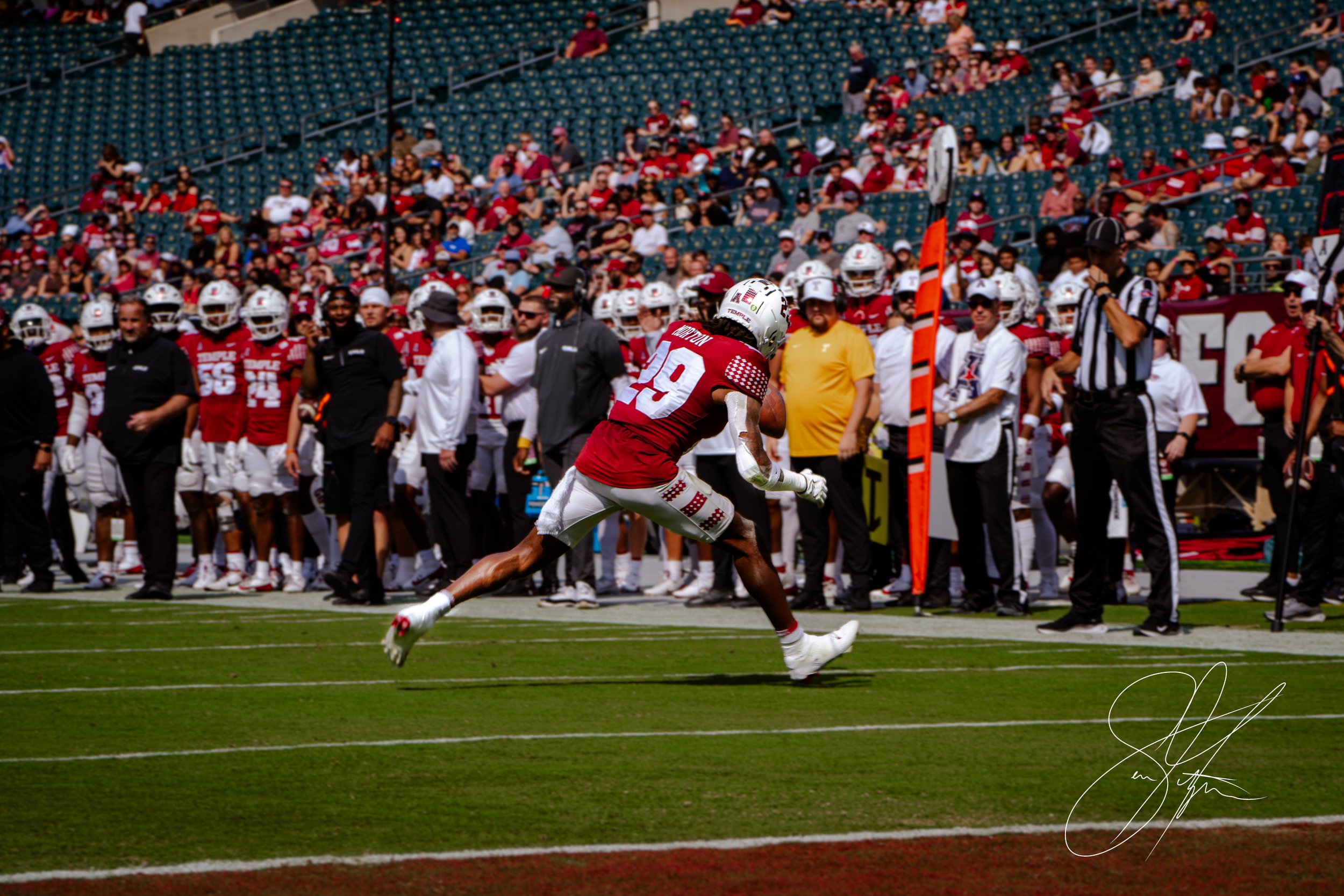 Football player catching a pass during a game, wearing red and white uniform, with teammates and referee visible on the sideline and audience in the background.