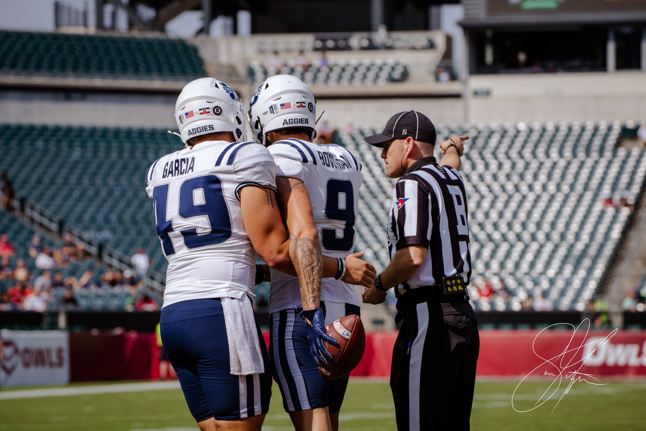Two football players in blue and white uniforms stand with a referee pointing on the sidelines of a stadium.