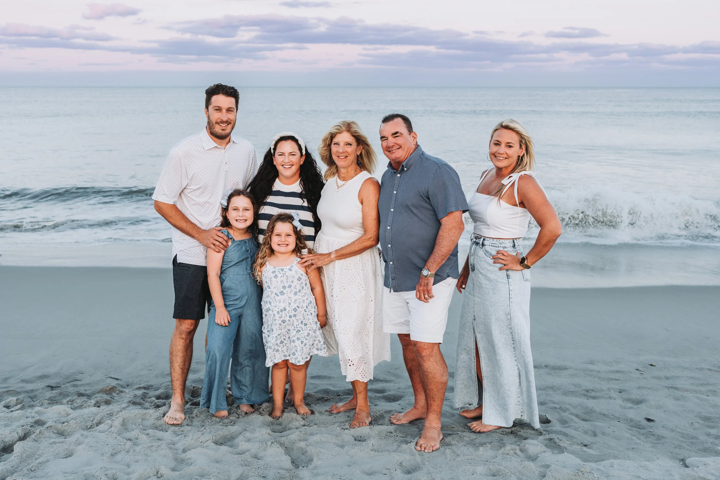 A family of seven posing on a beach with the ocean and cloudy sky in the background. Two young girls are standing in front of five adults. The group is barefoot and dressed in casual summer clothing.