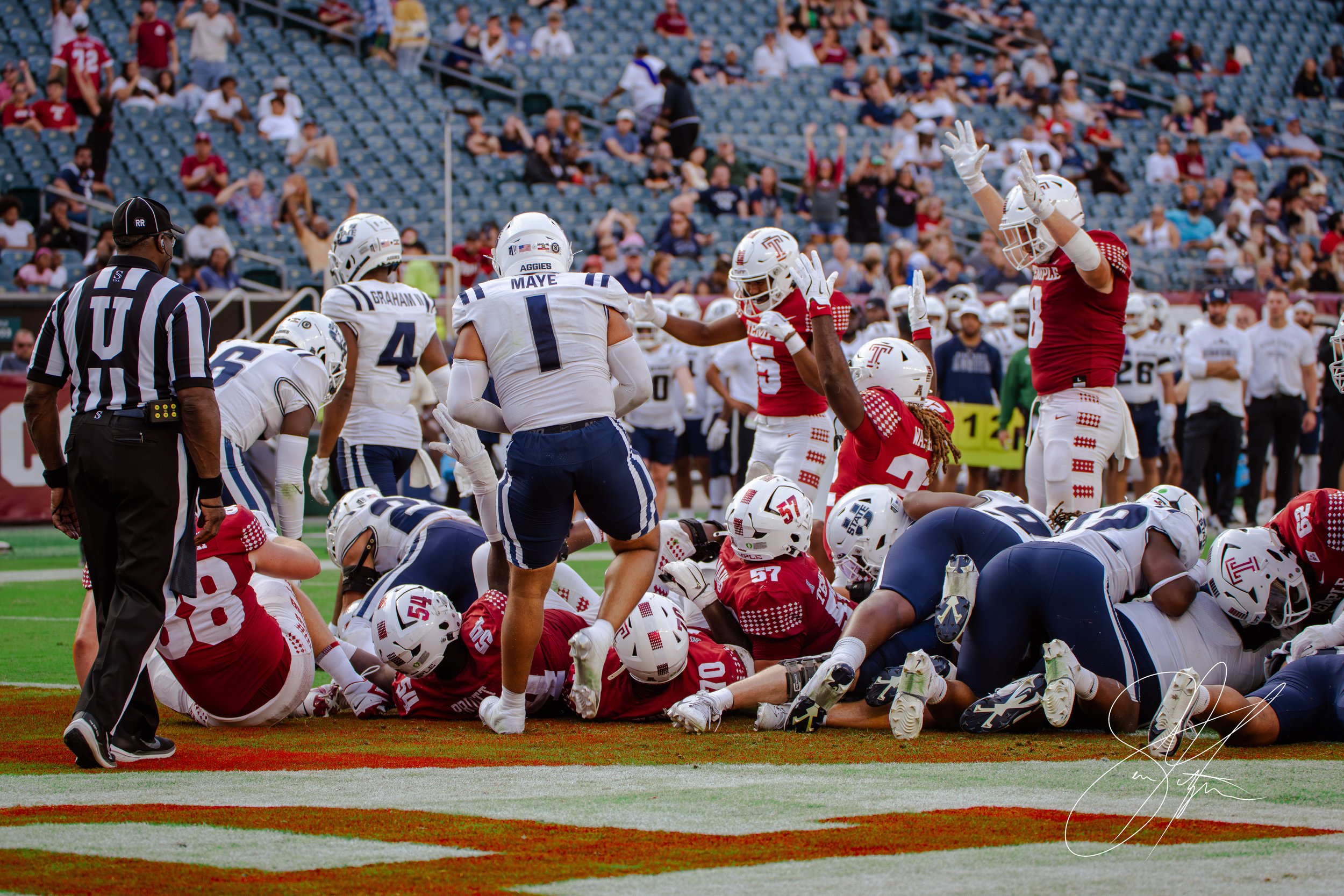 Football players from two teams, one in red and the other in white and blue, are piled on the field during a game. A referee stands nearby, and a player in red raises his arms, with spectators in the background.