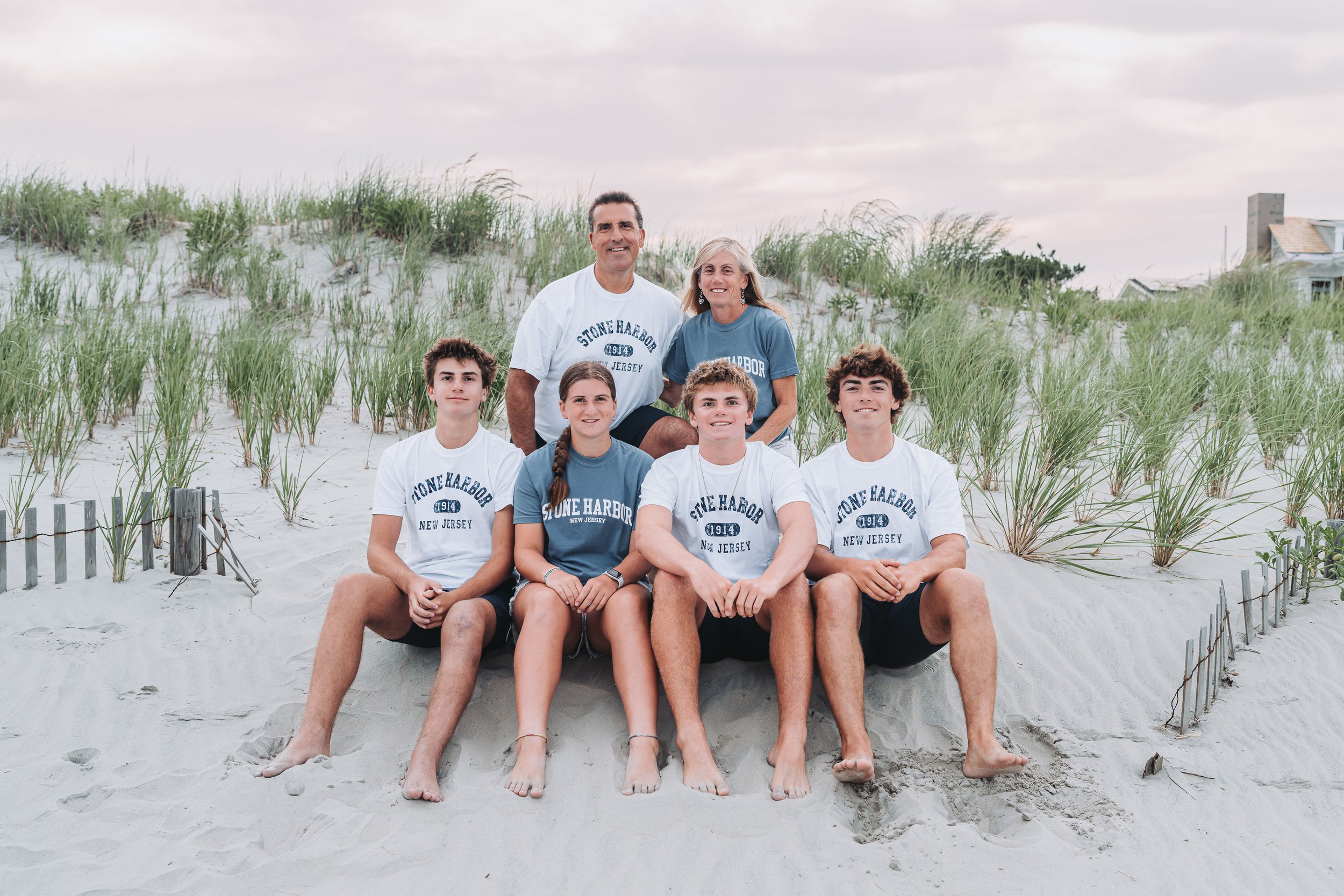 Family of six sitting on sandy beach with dune grass, wearing matching "Stone Harbor New Jersey" shirts.