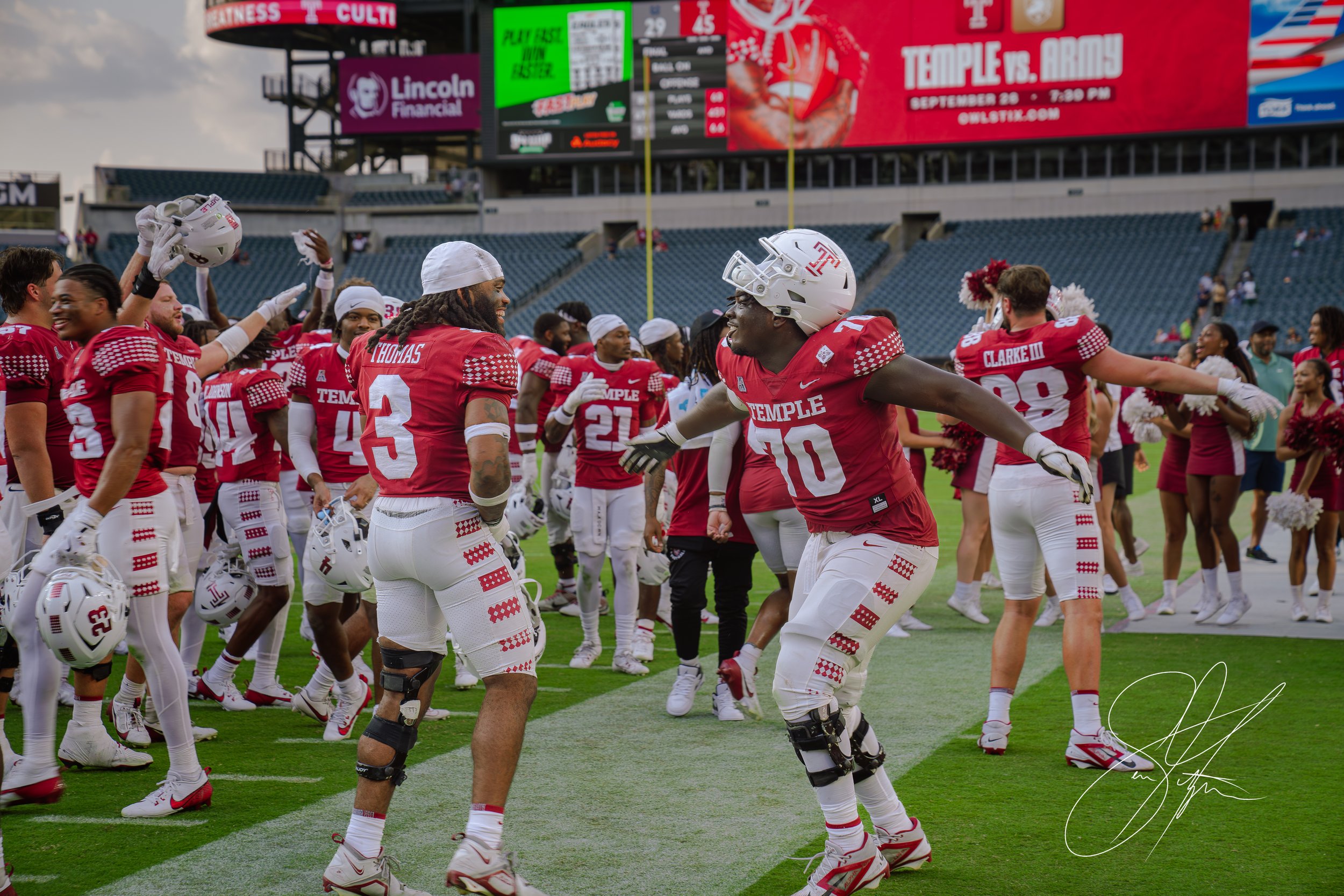 Temple University football players in red uniforms celebrating on the field. A scoreboard and cheerleaders are in the background.