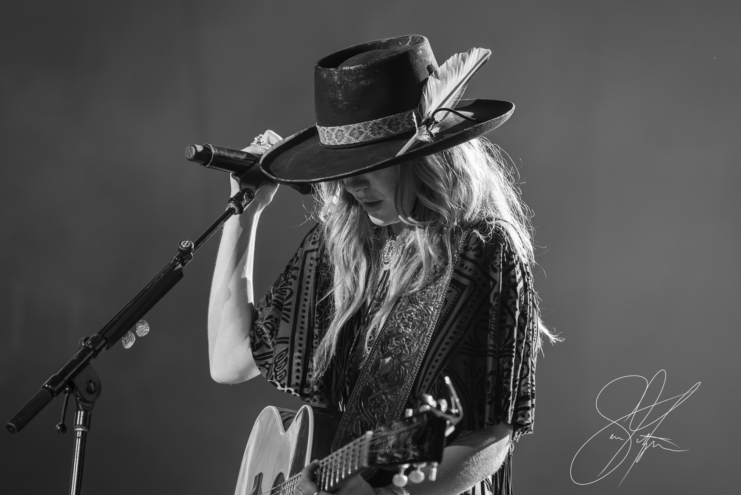 Black and white photo of a musician wearing a wide-brimmed hat with a feather, holding a microphone, and playing an acoustic guitar.
